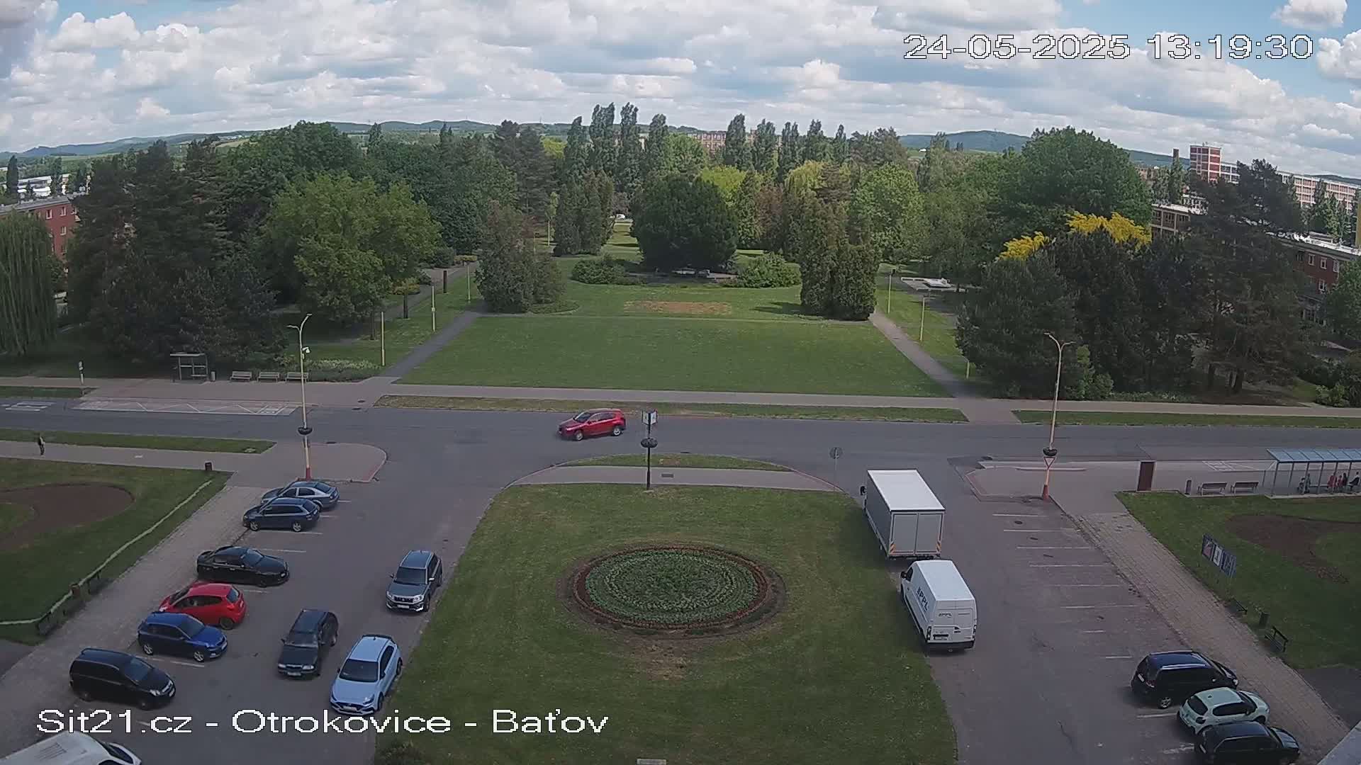 A high-angle view of a park-like area with a large grassy field, several cars parked in a lot, and two white delivery trucks, all under a partly cloudy sky.