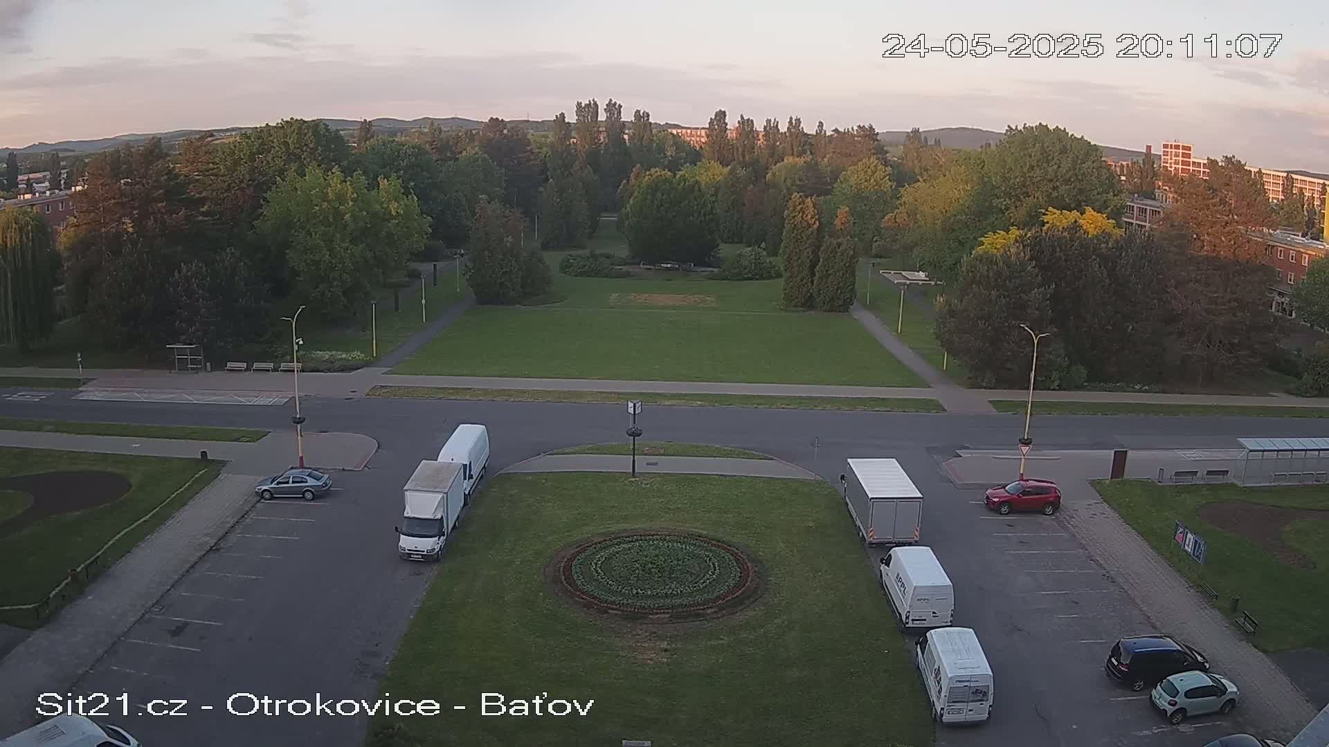 A high-angle, twilight view of a park with several vehicles parked in a lot, surrounded by buildings and trees.