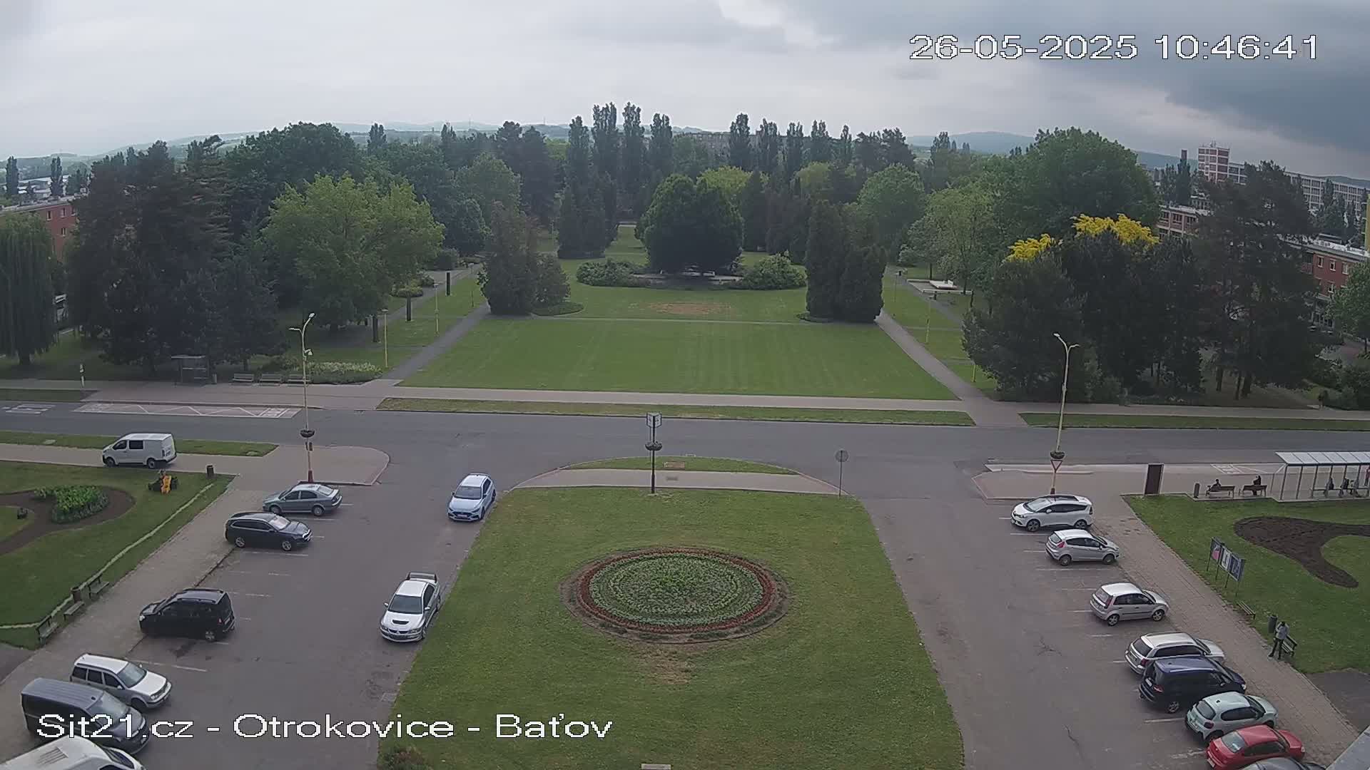 An overhead view shows a park with a large grassy area, surrounded by trees and buildings, under an overcast sky; several cars are parked in a lot nearby.