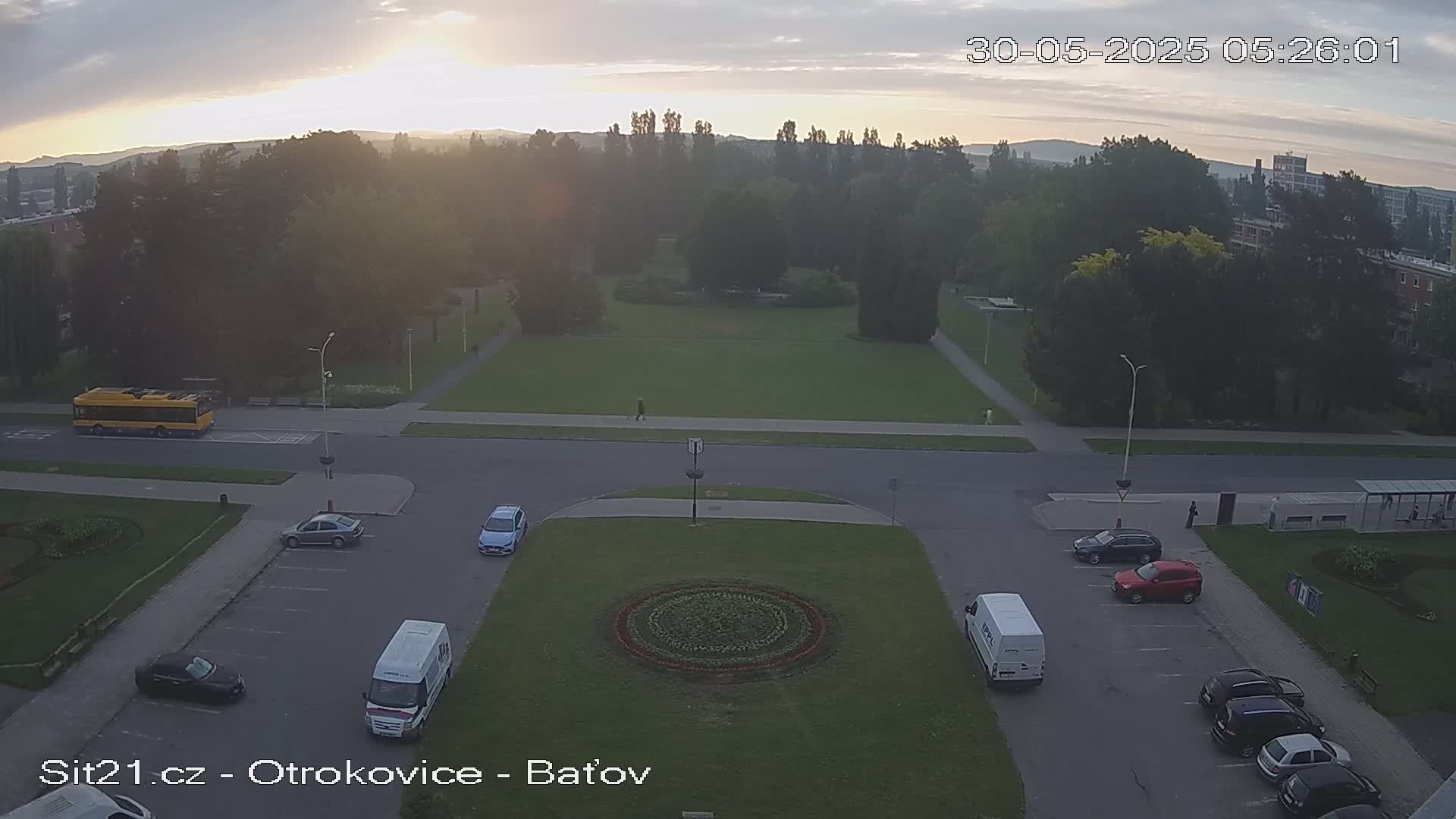 A high-angle, long shot shows a sunrise over a town square with a grassy area, several parked cars, a bus, and a van.