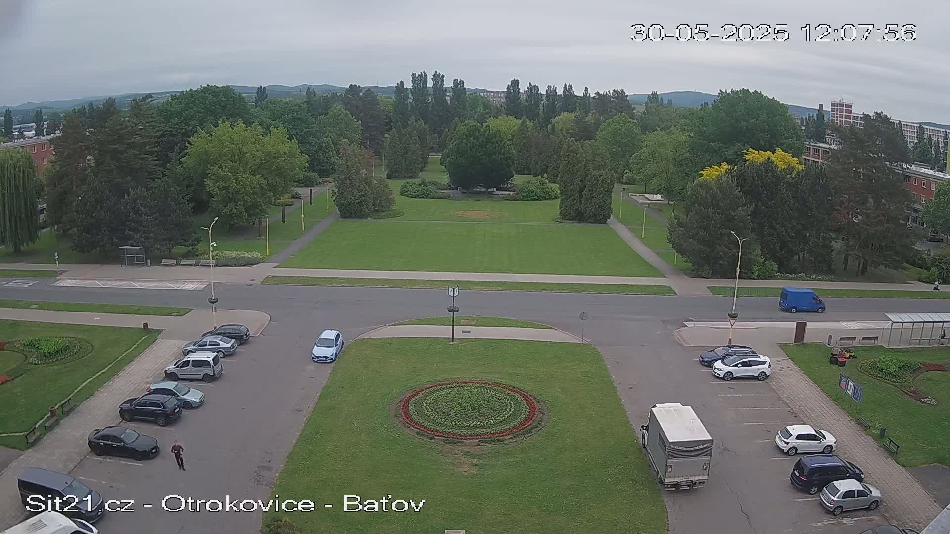 An elevated view shows a park with a large grassy area, surrounded by trees and buildings, under an overcast sky; cars are parked in a lot nearby.
