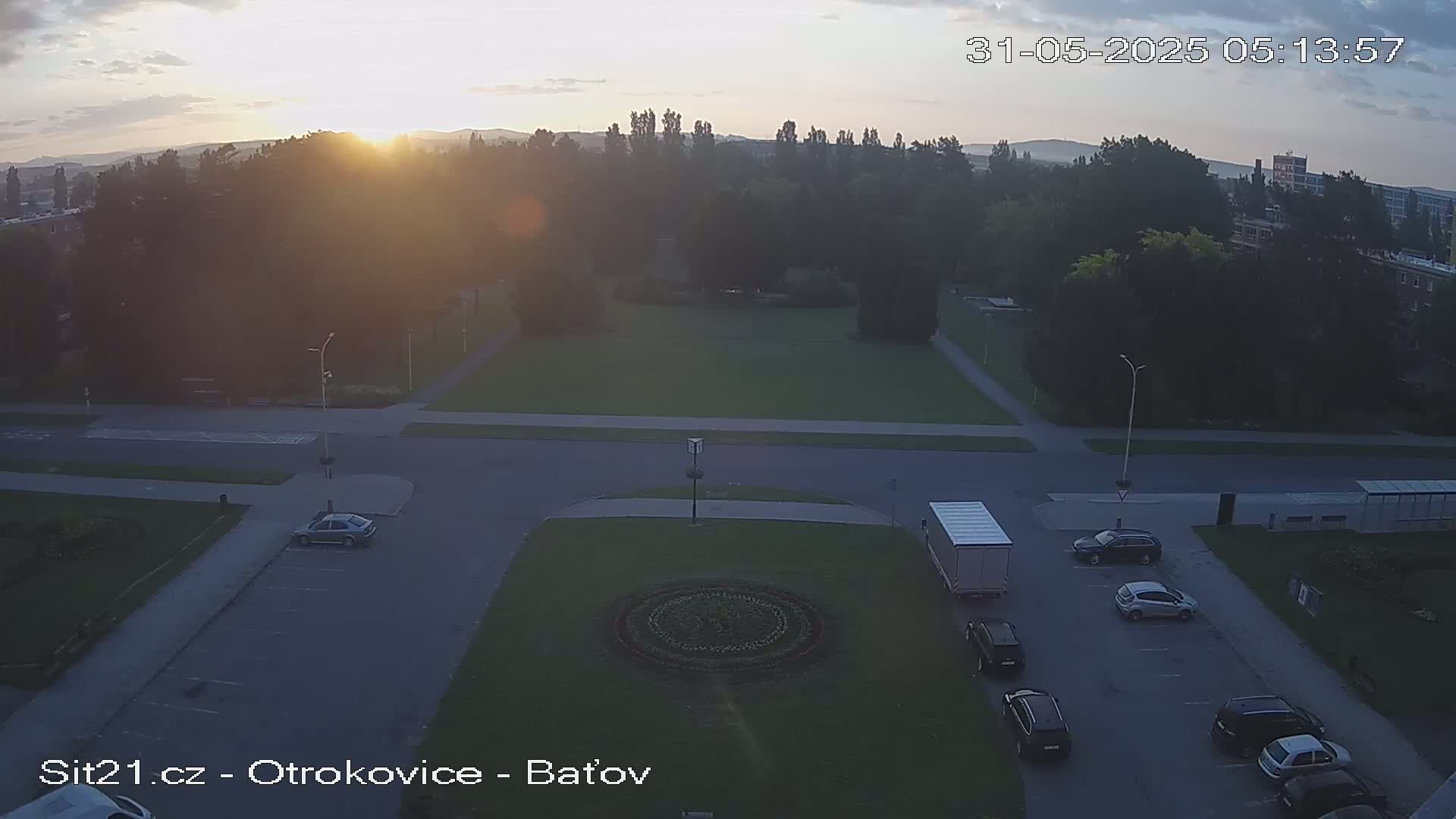 A high-angle, long shot at sunrise shows a park with a circular flower bed in the center, surrounded by a parking lot with several cars and a truck; the weather is clear.