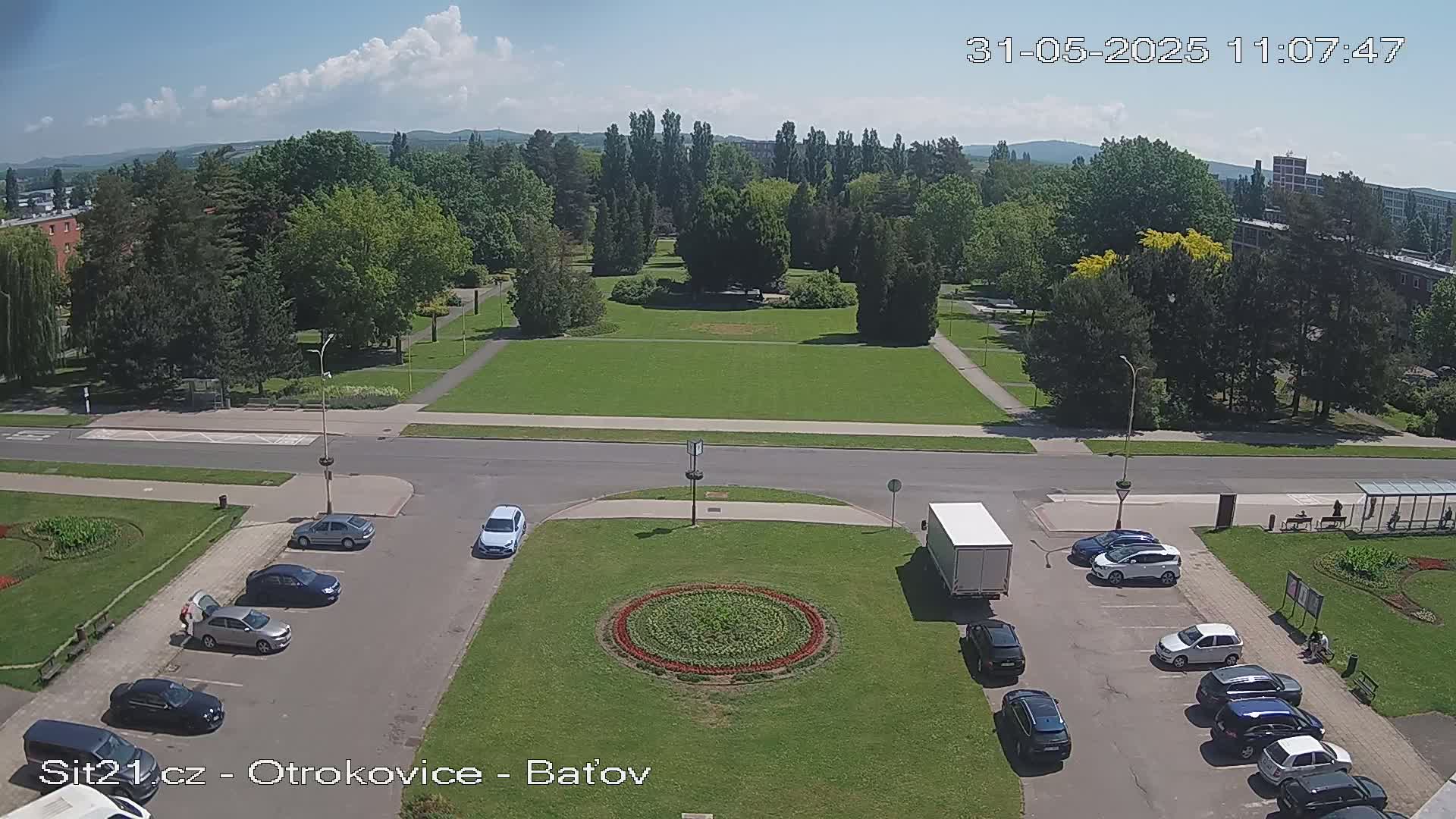 A sunny day shows a large grassy park surrounded by trees and buildings, with several cars parked in a lot next to a circular flower bed.