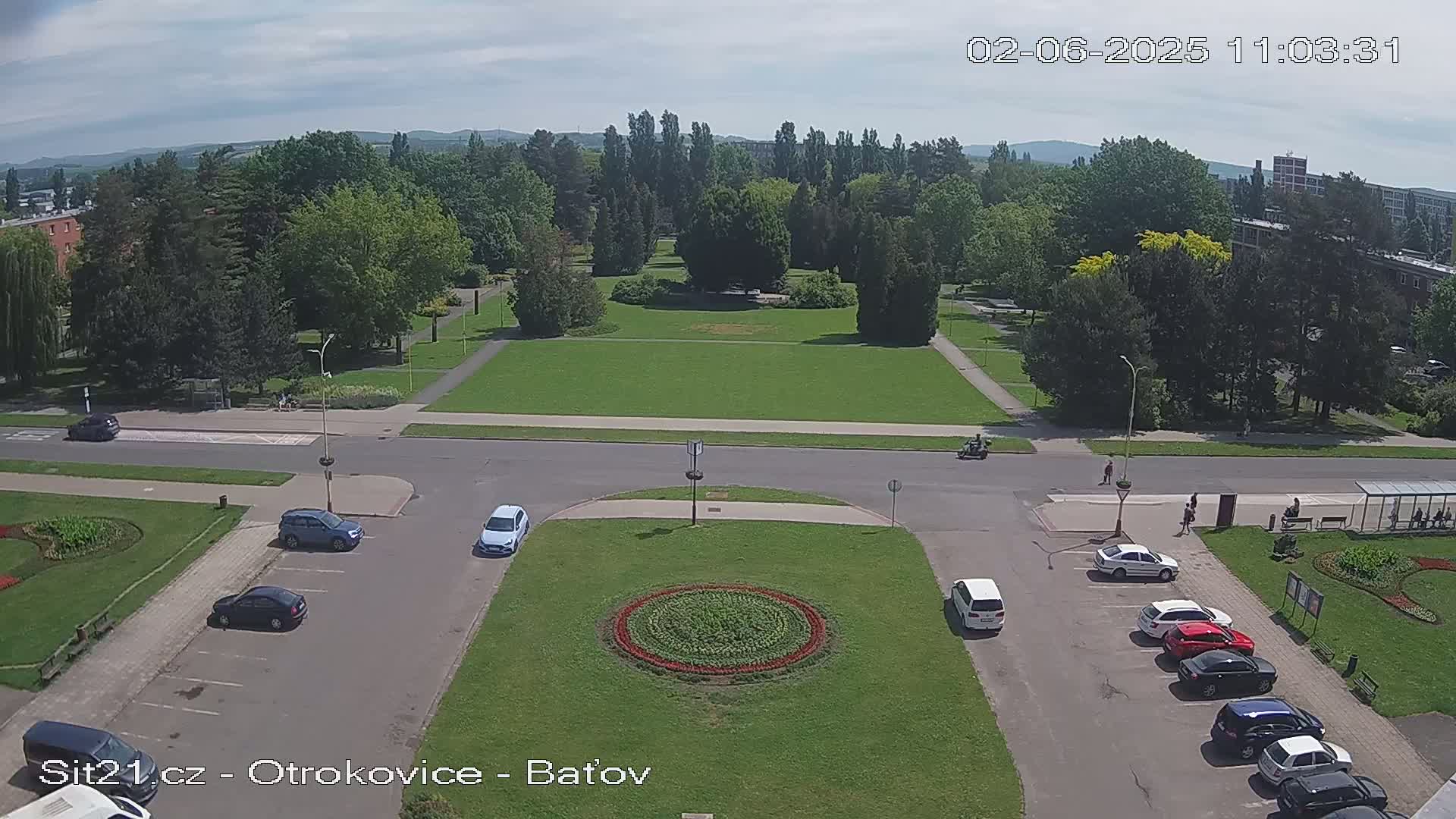 A high-angle view of a park with a large grassy area and several cars parked in a lot under a partly sunny sky.