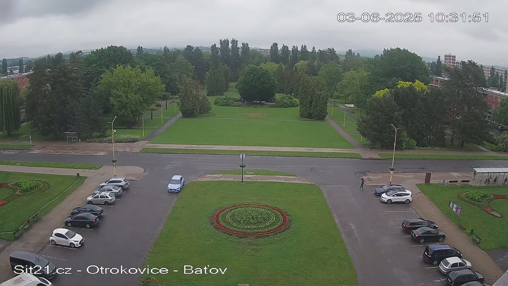 A high-angle view shows a park with a circular flower bed in the center of a roadway, surrounded by parked cars and overcast skies.