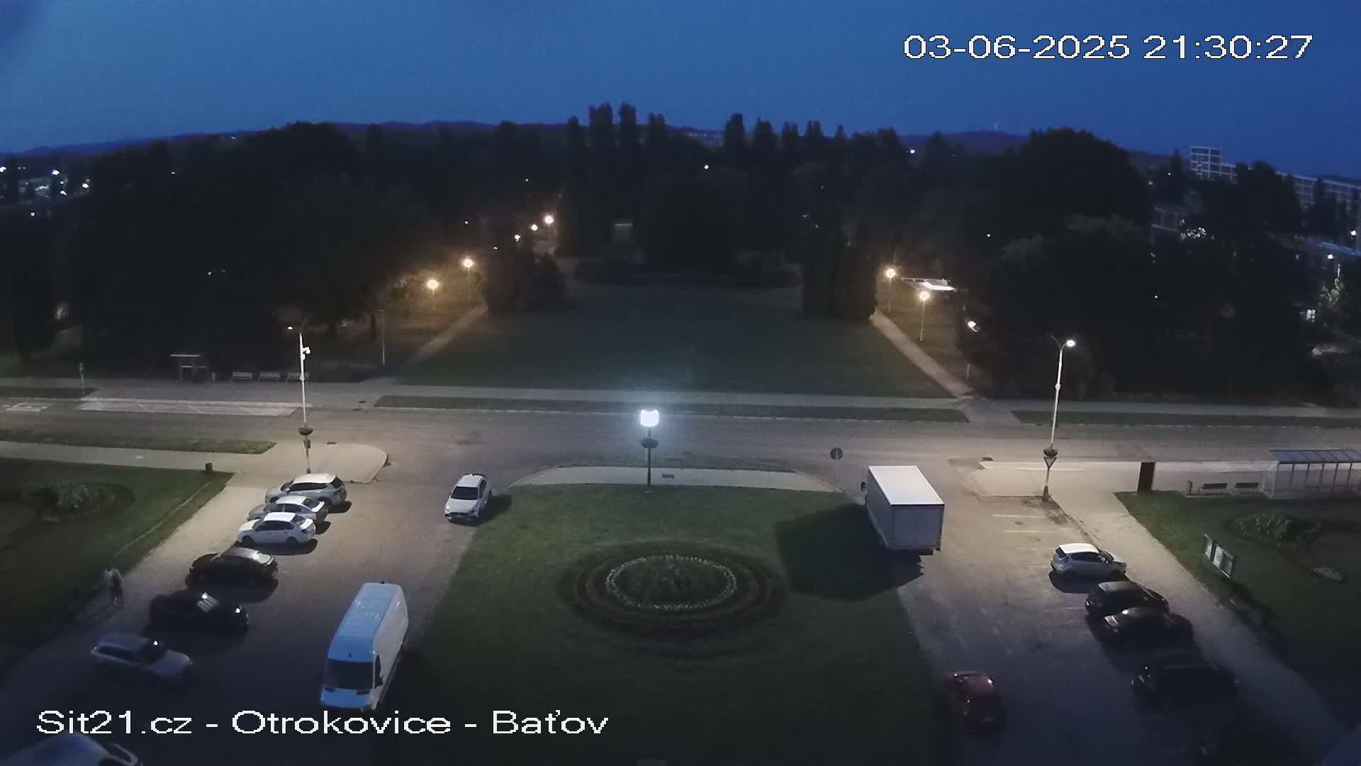 A nighttime aerial view shows a town square with cars parked, a truck, and a circular flower bed in the center, under a dark blue, twilight sky.
