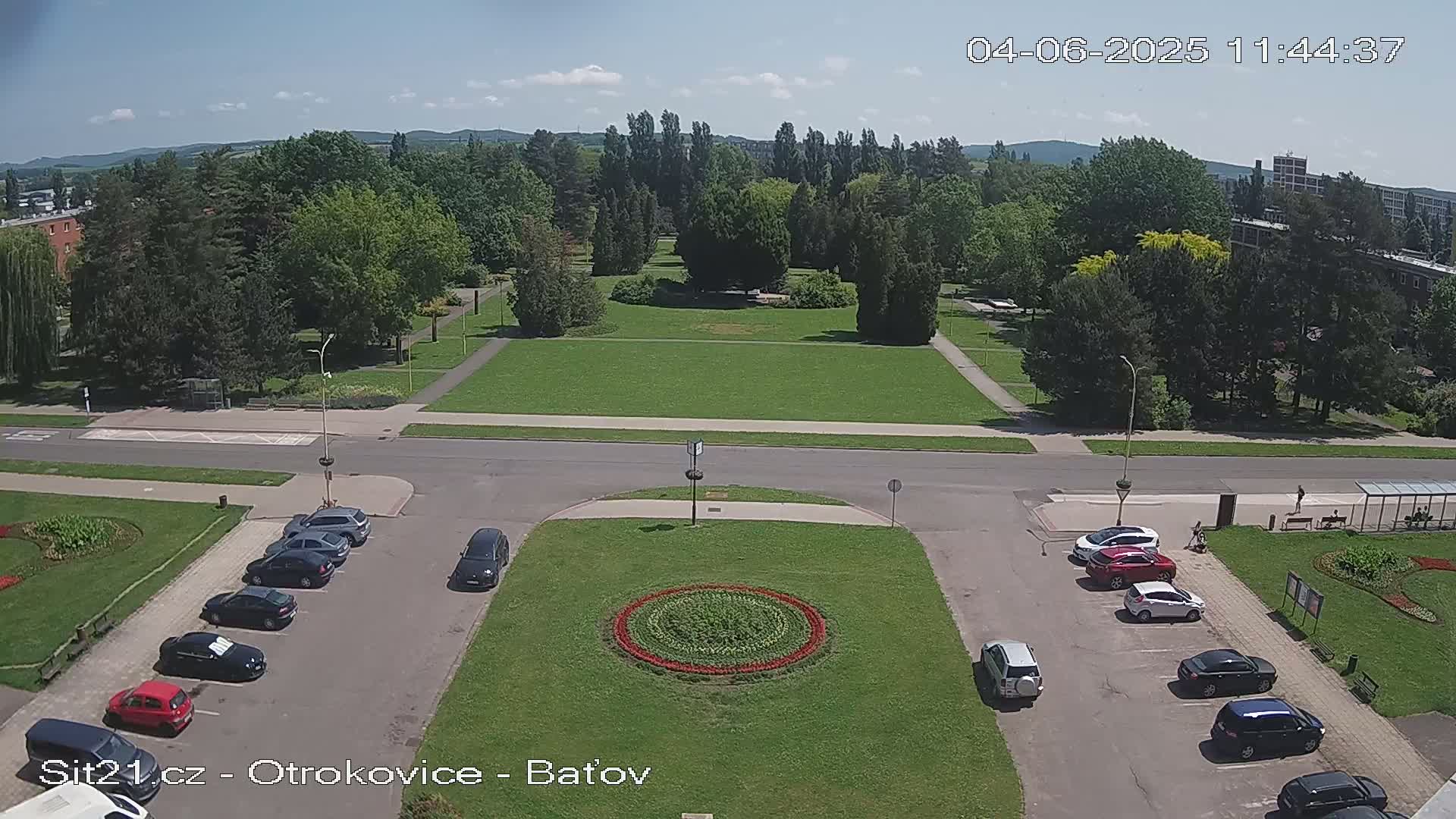 A sunny day reveals a parking lot with several cars surrounding a circular flower bed in a grassy area, across a road from a large park with lush green trees and lawns.