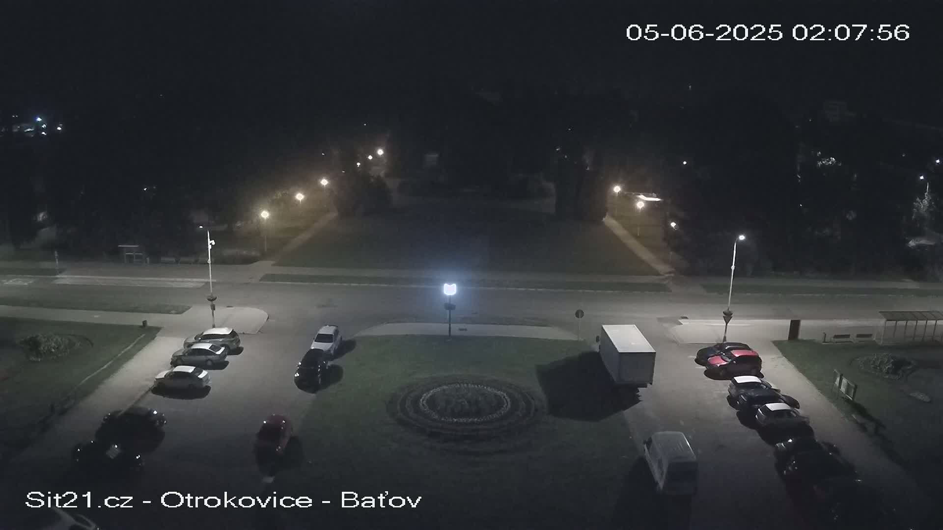 At night, several cars are parked in a lot surrounding a circular garden, with streetlights illuminating the scene.