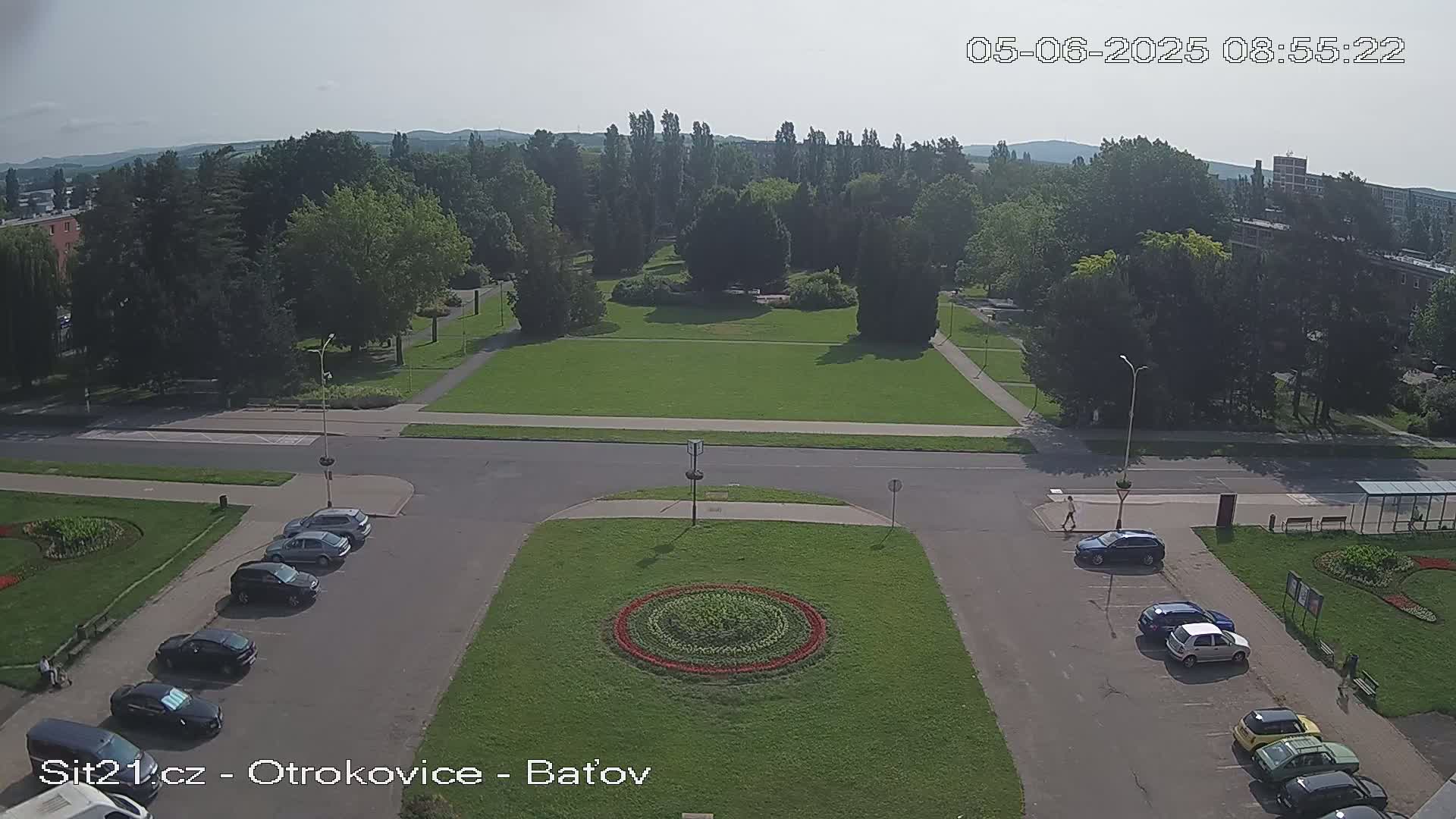 An elevated view of a park and surrounding area on a sunny day, showing cars parked in a lot near a roadway and a large circular flower bed in the center of a grassy area.