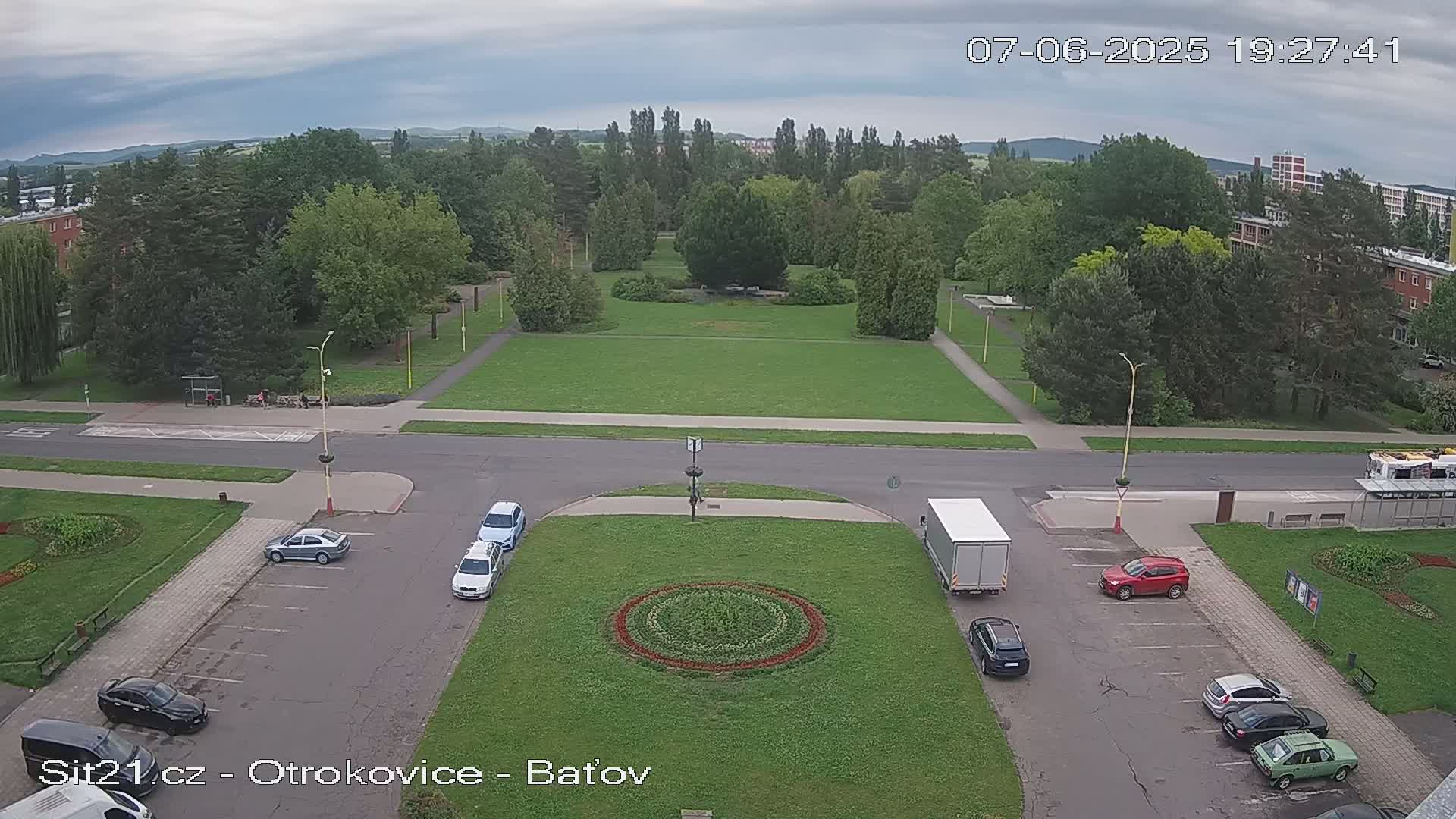 A high-angle view shows a park with a circular flower bed in the center, surrounded by a parking lot with several cars, and a street leading to a larger park and buildings, under a cloudy sky.