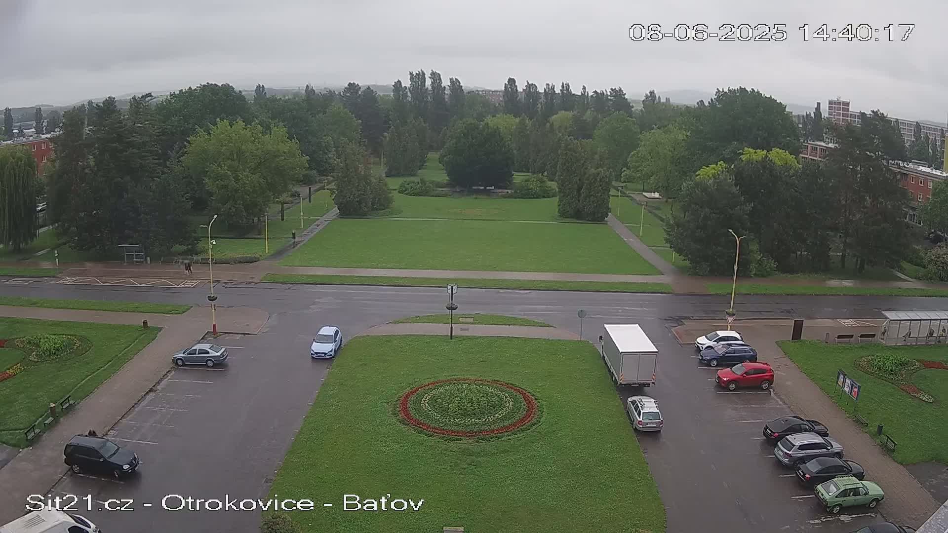 A high-angle view of a town square on a cloudy day, showing a grassy park with a circular flower bed in the center, surrounded by parked cars and a street.
