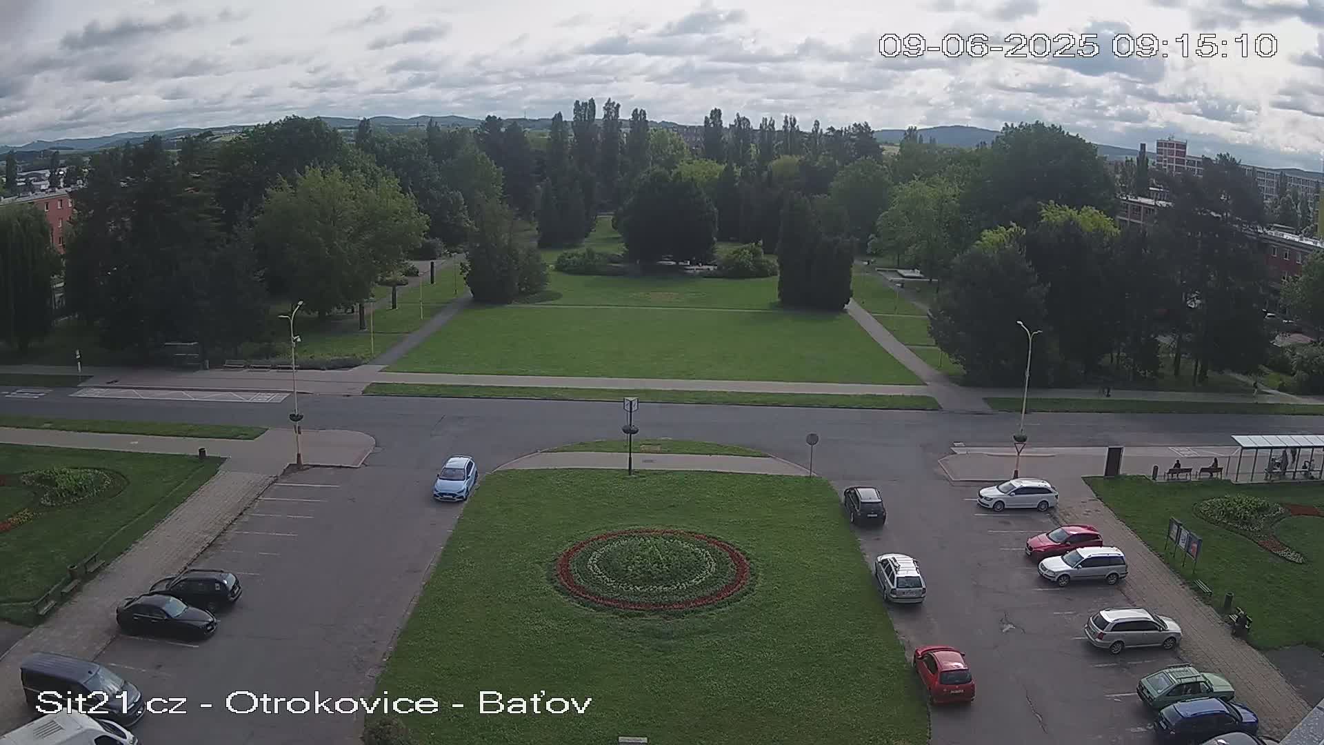 A large grassy park with several cars parked in a lot surrounding a circular flower bed, under a cloudy sky.