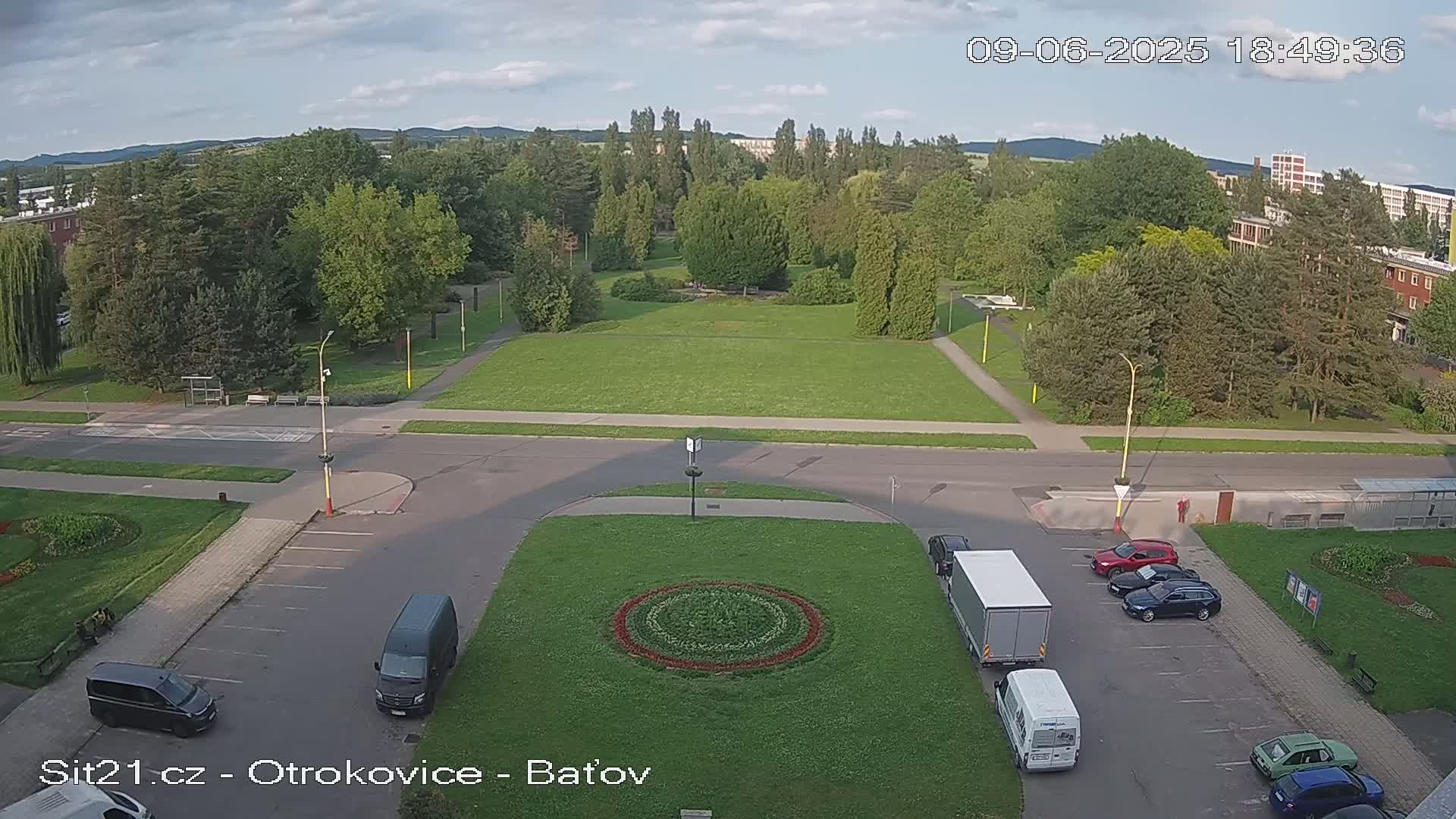 A mostly sunny aerial view of a park with a circular flower bed in the center of a parking lot, surrounded by roads, buildings, and parked cars.