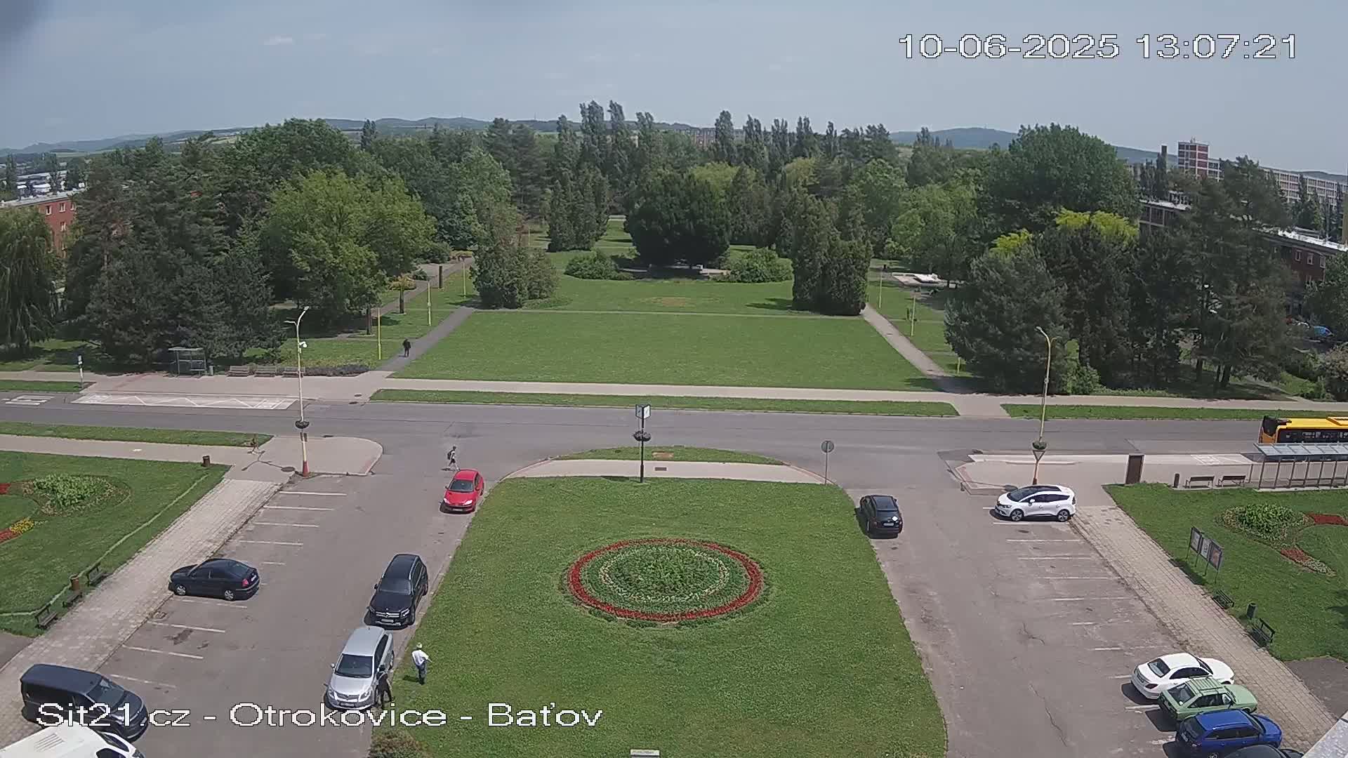 A high-angle view of a park and surrounding area on a sunny day, showing a road, parking lot with several cars, and a circular flower bed in the center of a grassy area.