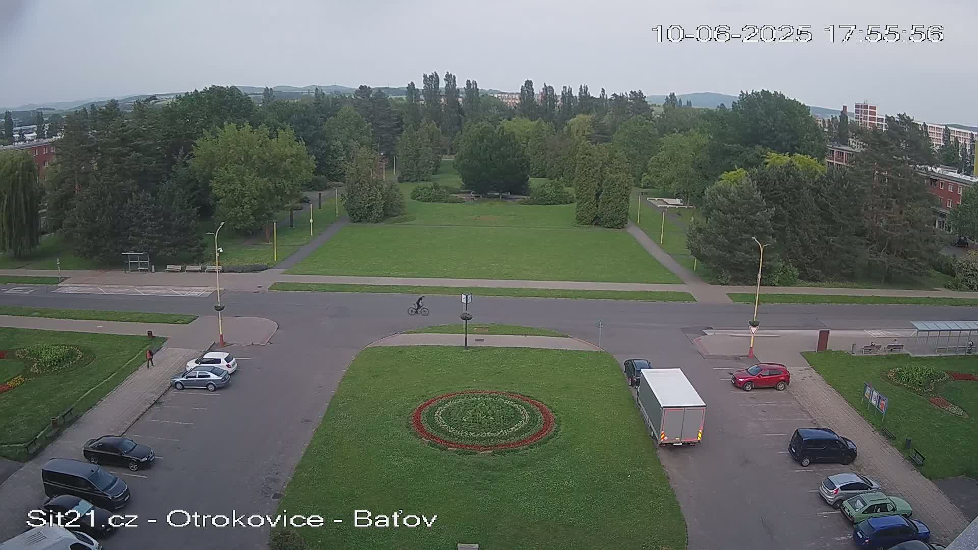 An elevated view of a park and parking lot with several cars, a bicycle rider, and a truck, under an overcast sky.