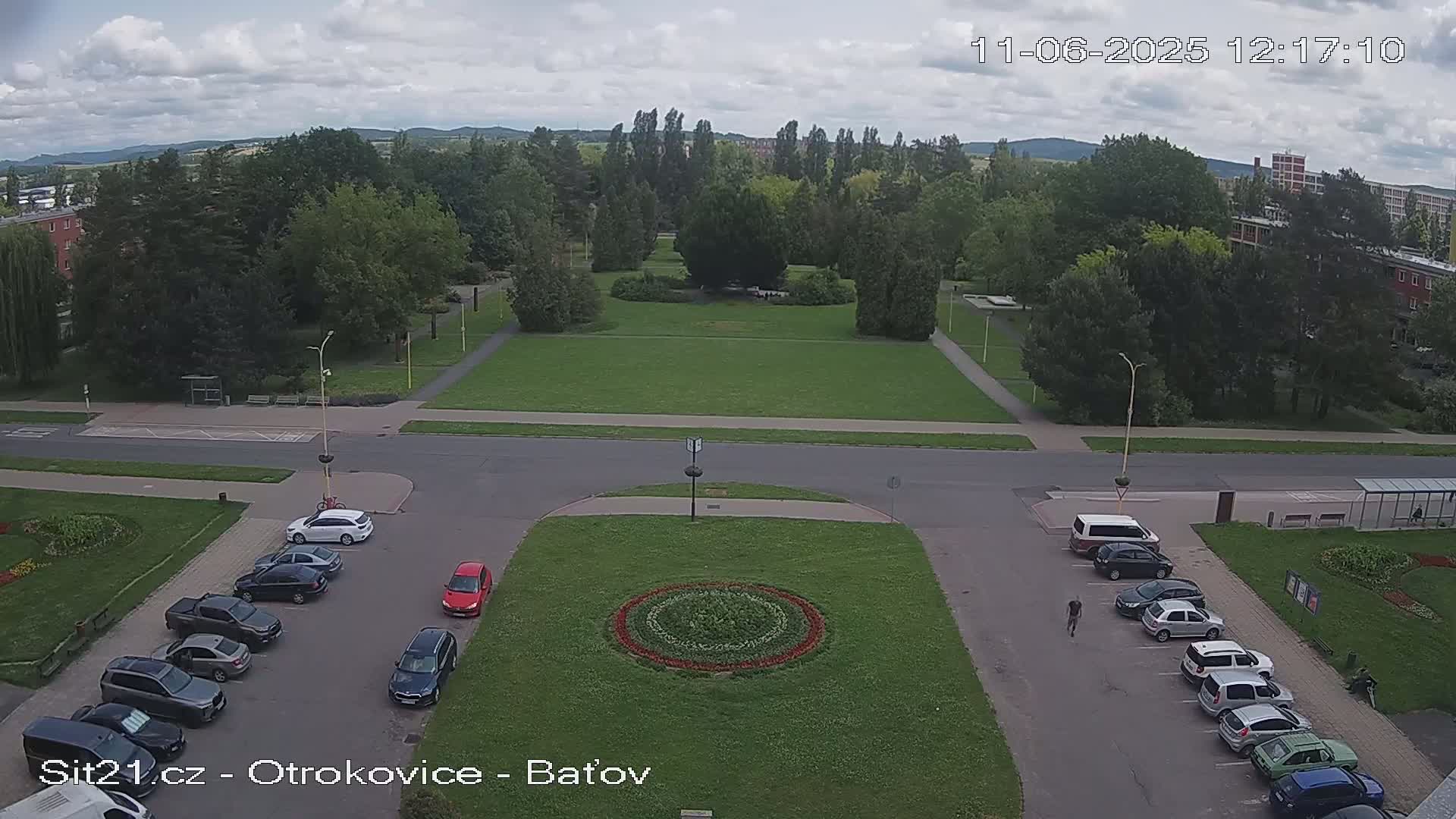An overhead view of a town square with a circular flower bed, surrounded by parked cars and trees under a partly cloudy sky.
