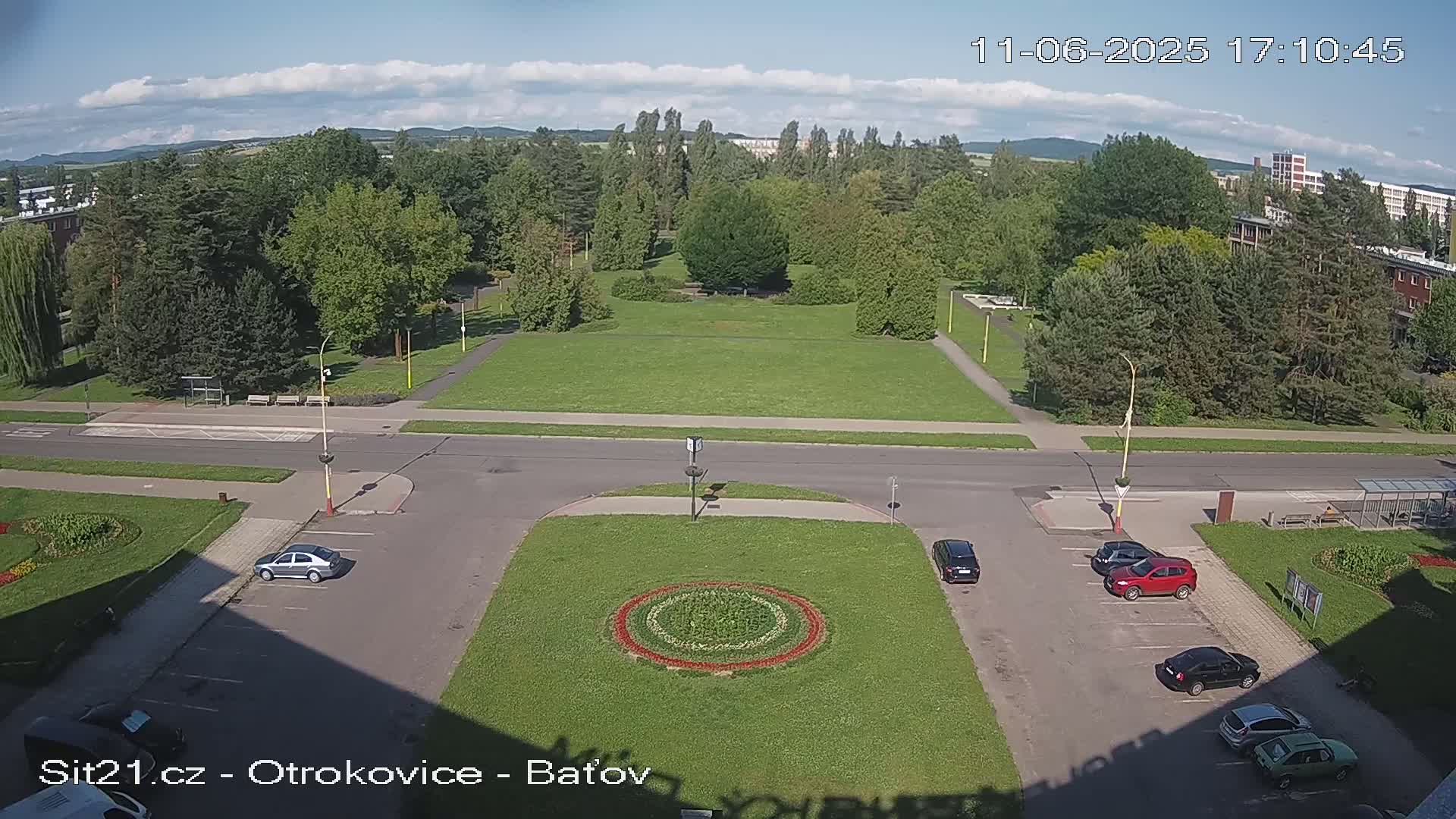 A high-angle view of a sunny parking lot and a grassy park surrounded by buildings and trees.