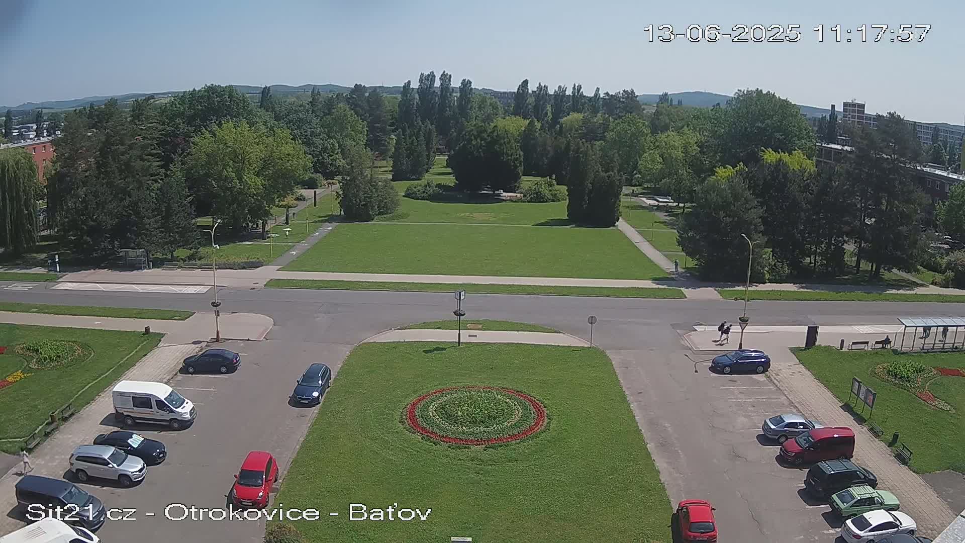 A high-angle view of a park and parking lot on a sunny day, with several cars parked and a few people walking.