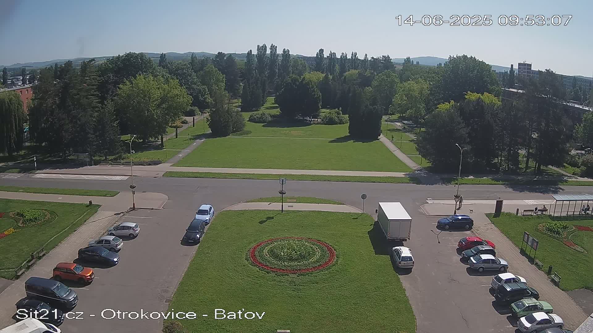 A high-angle, sunny view shows a town square with a circular flower bed in the center, surrounded by parked cars and a grassy park beyond a roadway.