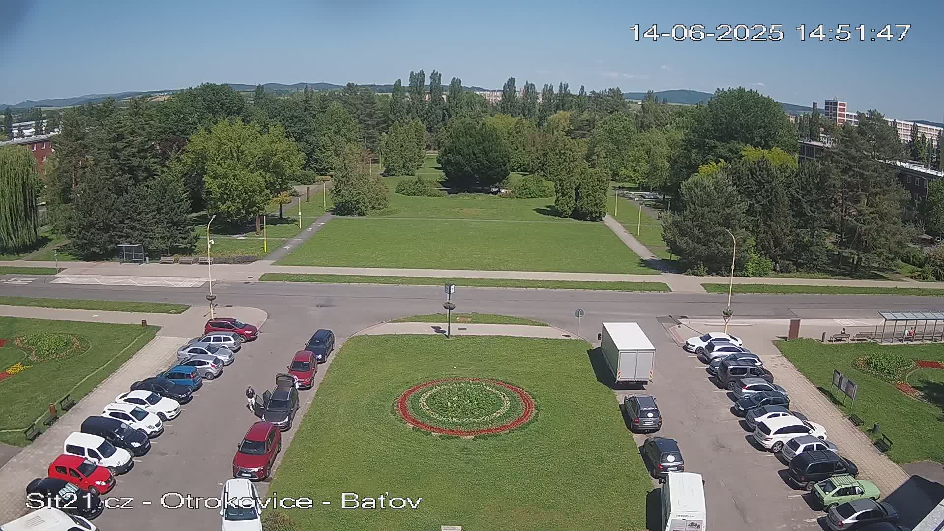 An aerial view shows a sunny day with cars parked along a road next to a green grassy park with a circular flower bed in the center.