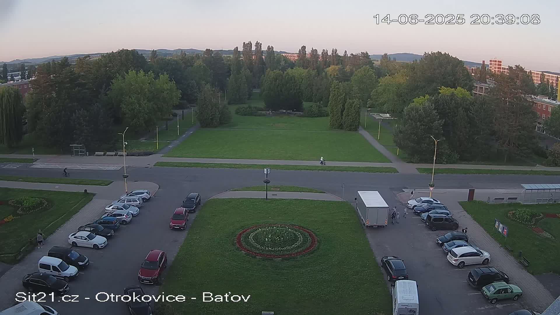 An aerial view shows a sunny day with cars parked along a road next to a green grassy park with a circular flower bed in the center.