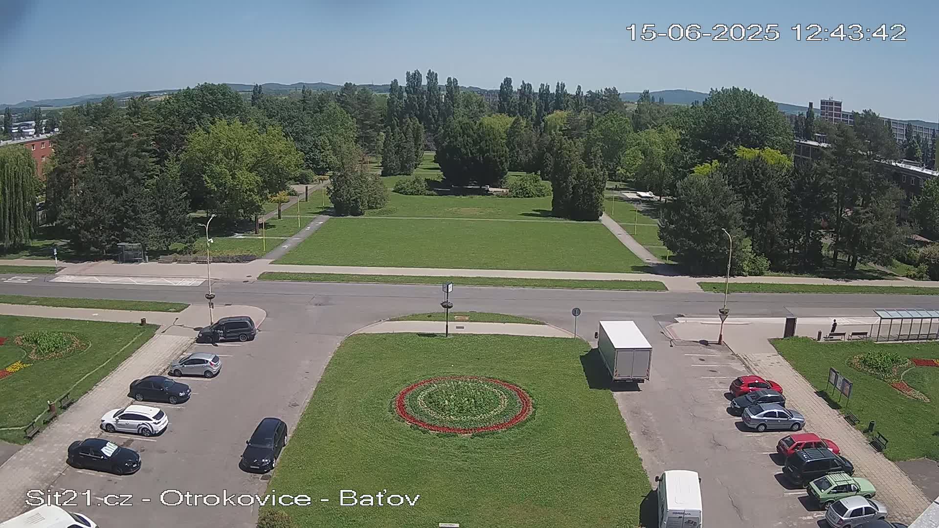 A sunny day shows a parking lot with several cars and a truck, next to a park with a circular flower bed, all bordered by a road and green trees.