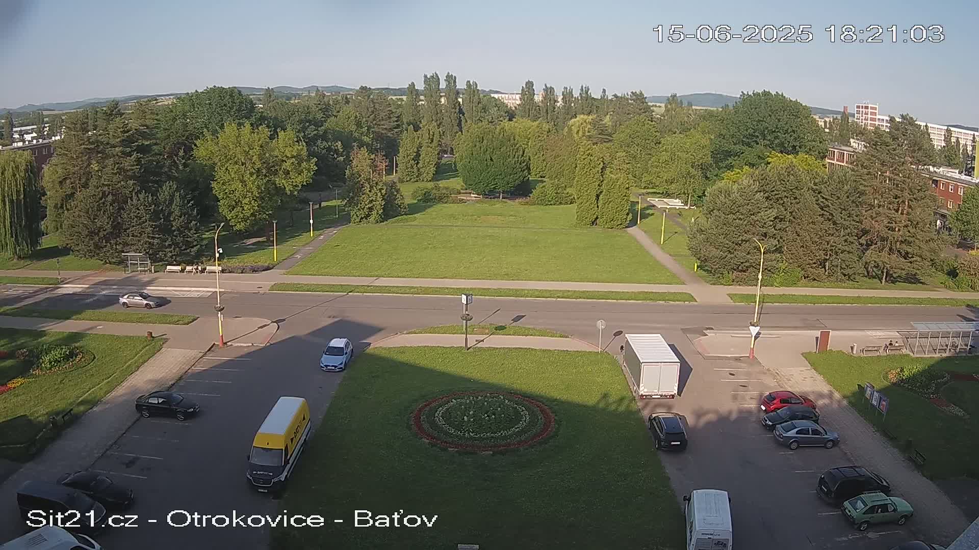 An aerial view shows a sunny day over a park with several vehicles parked in a lot near a road.