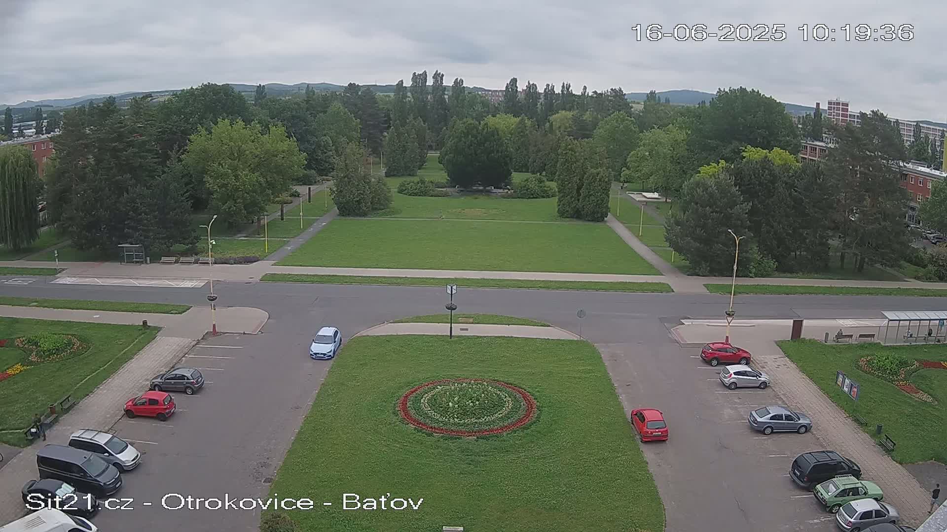 A parking lot with several cars, a grassy area with a circular flower bed, and a larger park with trees under an overcast sky.