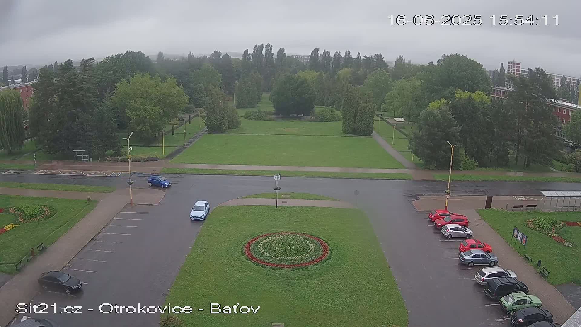 A high-angle view of a park with a circular flower bed in the center, surrounded by a parking lot with several cars, under an overcast sky during rain.