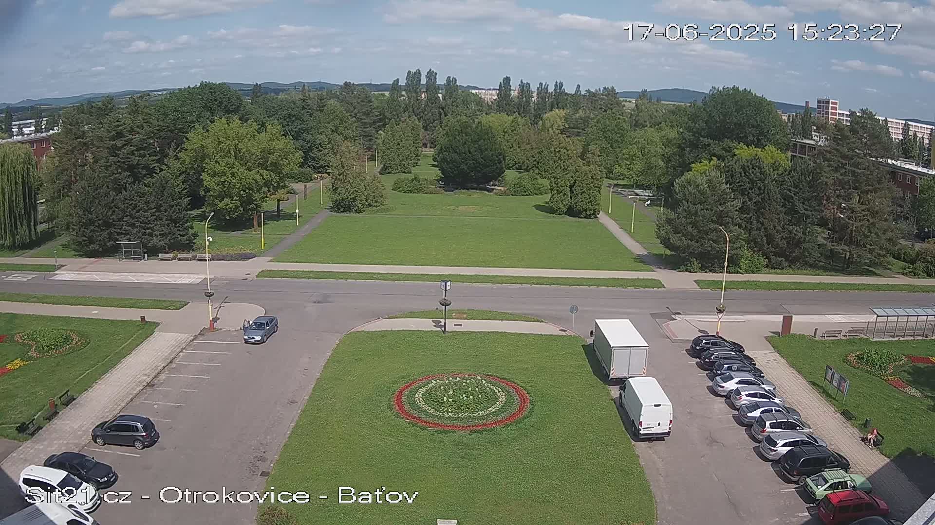 A high-angle view shows a parking lot with several cars and a truck, next to a grassy area with a circular flower bed, a road, and a large park with many trees under a partly cloudy sky.