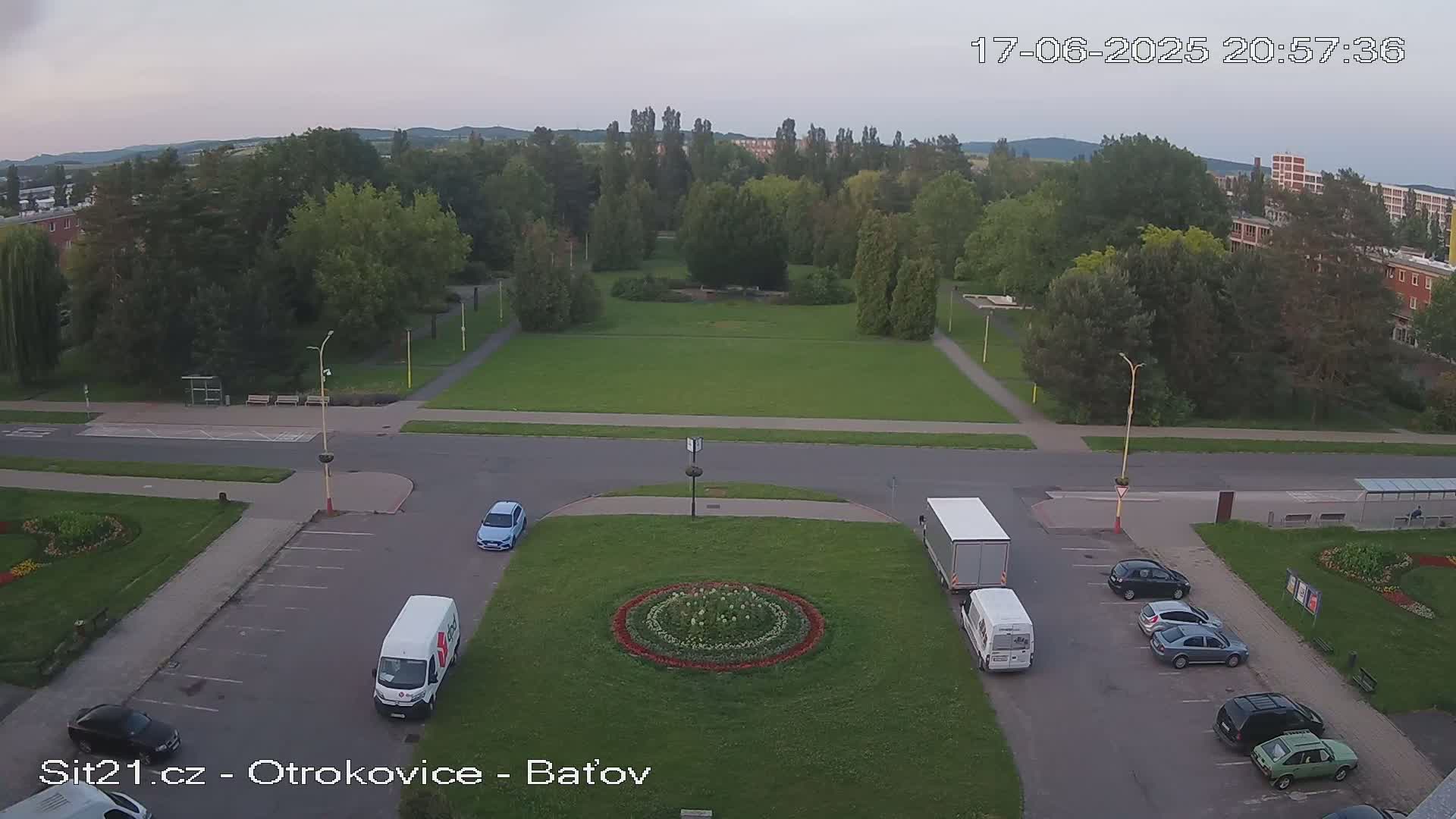 An aerial view of a town square with a circular flower bed at its center, several vehicles parked nearby, and a grassy park across the street, under a twilight sky.
