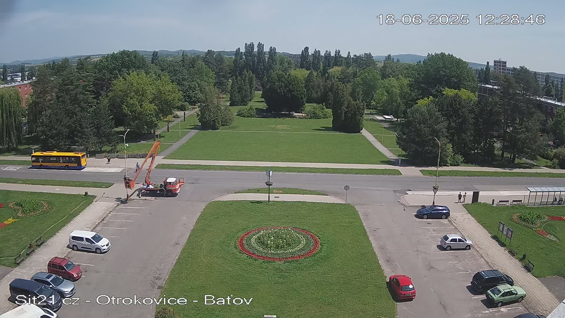 A high-angle, sunny-day view shows a large grassy area with a circular flower bed in the center, surrounded by a parking lot, a road, and a bus stop with a yellow bus.
