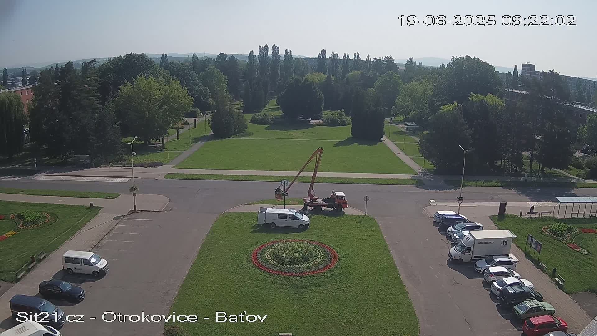 A high-angle, sunny-day view of a town square with a park in the background, featuring several vehicles parked along the perimeter and a cherry picker working near a circular flower bed in the center.