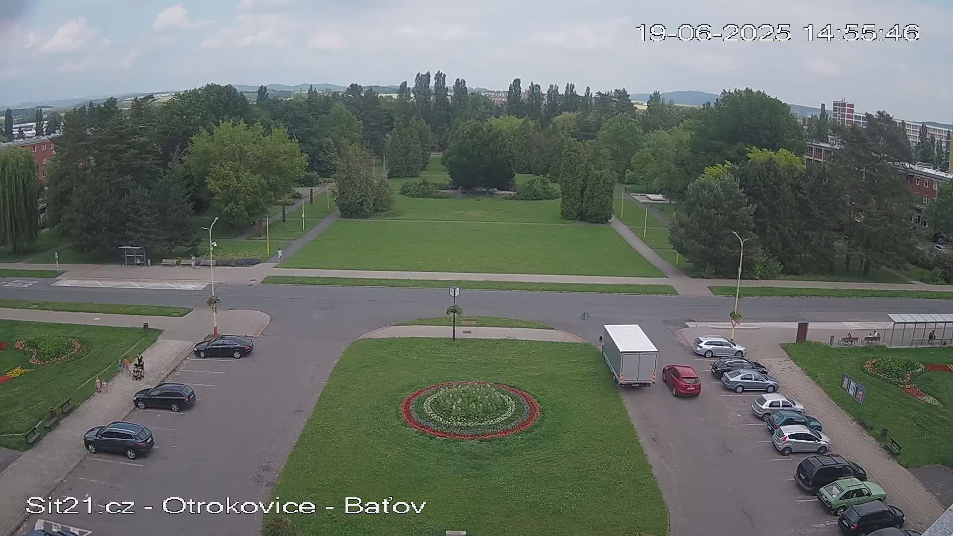A mostly sunny, overcast day reveals a large park with a circular flower bed in the center of a parking lot and several cars parked around it, surrounded by buildings and trees.