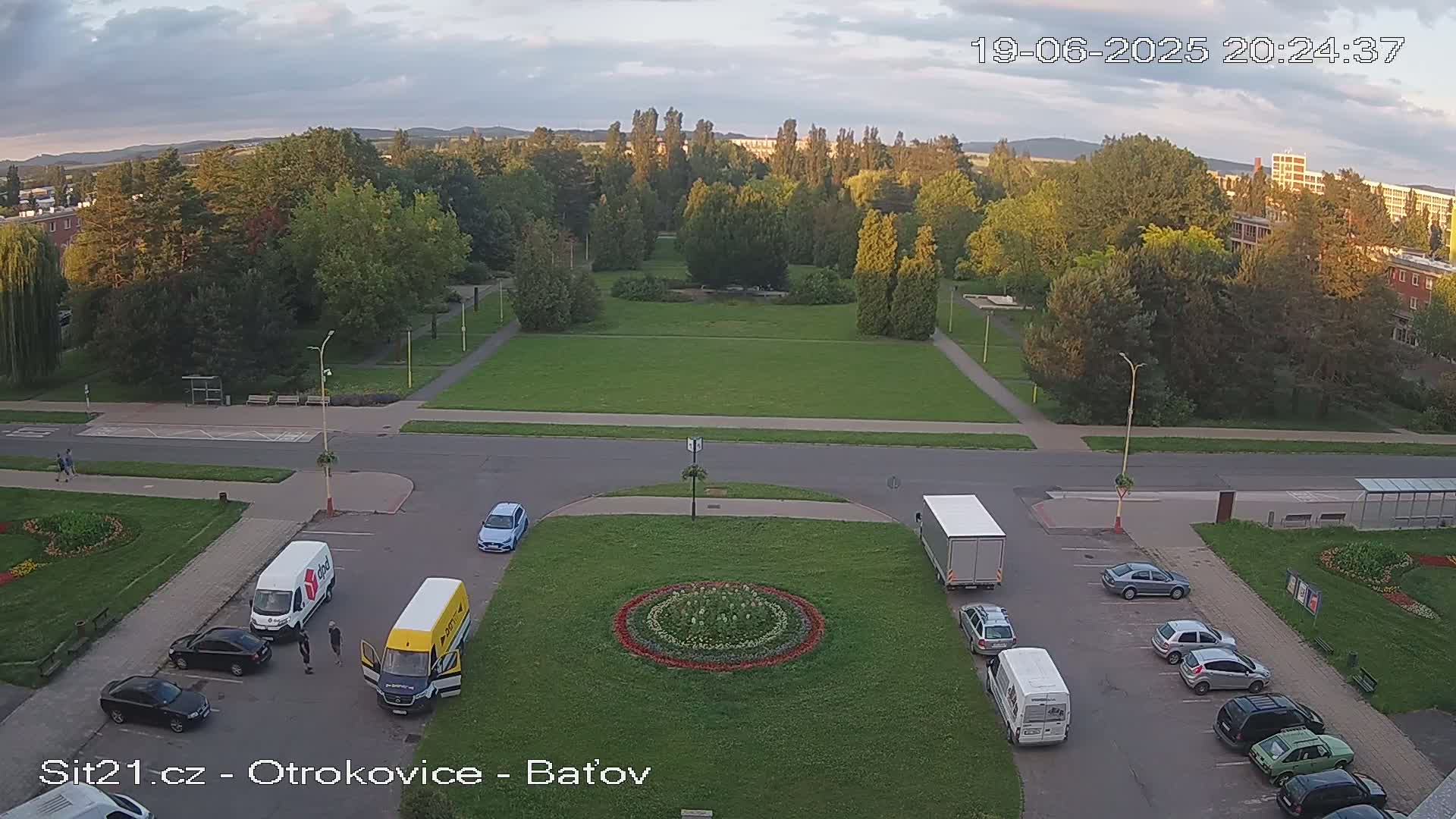 An aerial view of a park with a circular flower bed in the center, surrounded by a parking lot with several cars and vans, under a partly cloudy evening sky.