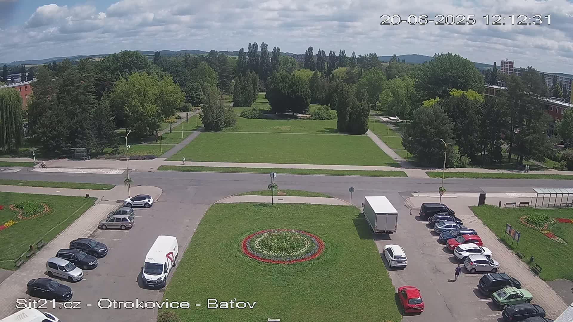 A mostly sunny day shows a large grassy park with several parked cars and a truck surrounding a circular flower bed in a town square.