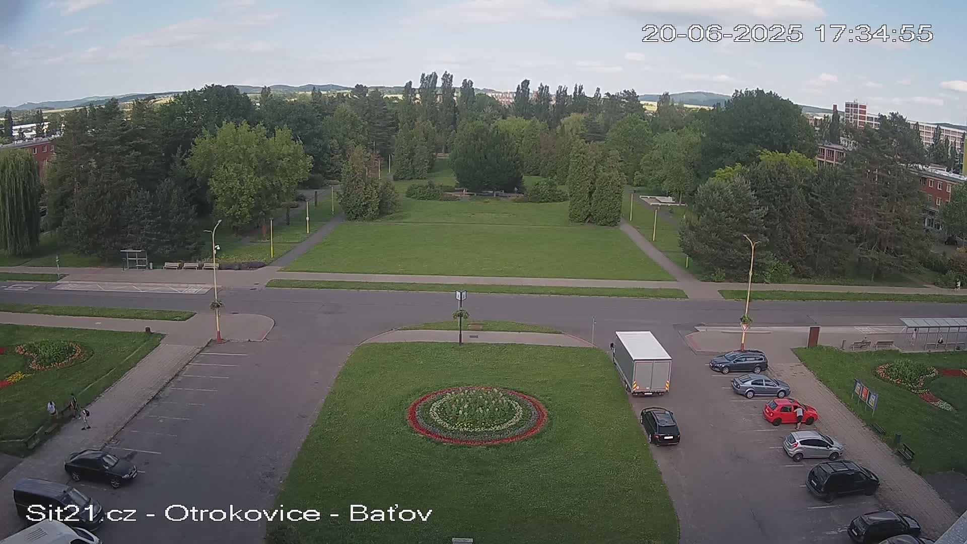 An aerial view shows a town square with a large grassy area, several parked cars, and a truck, under partly cloudy skies.