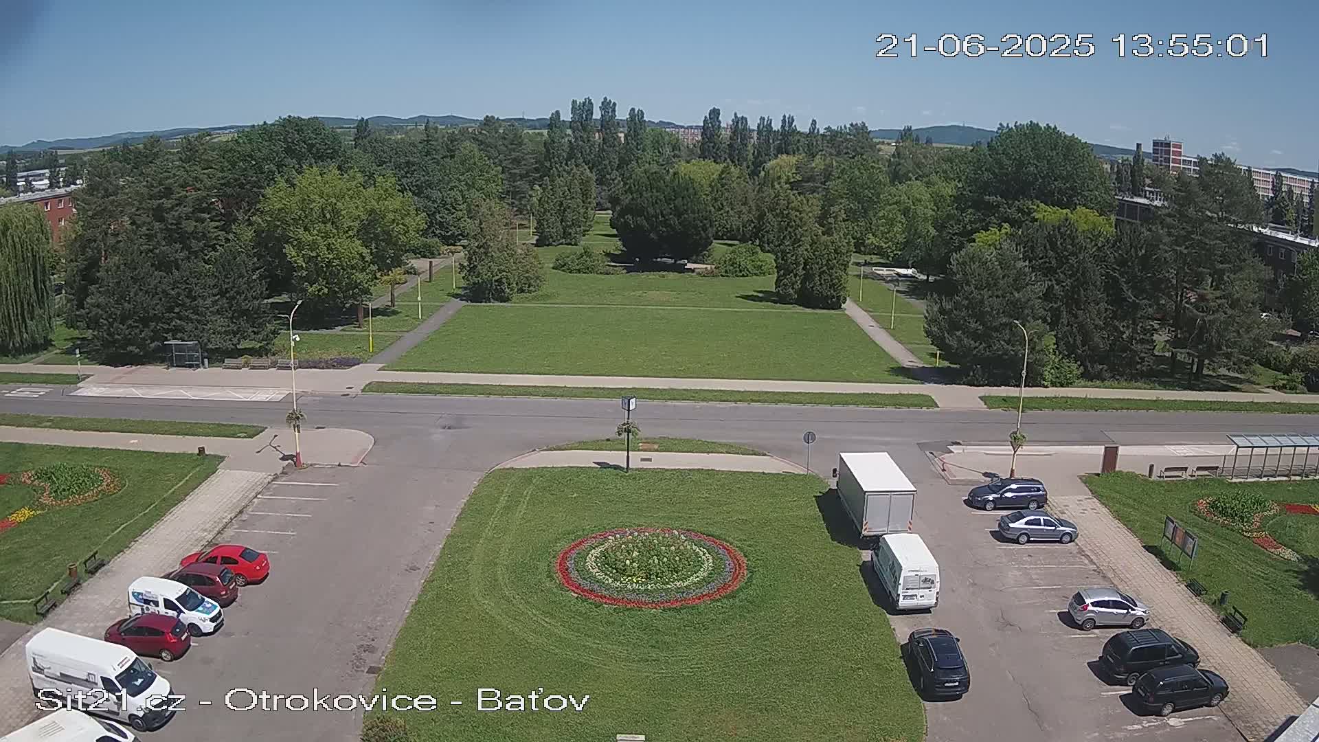 A sunny day reveals a park with a grassy expanse surrounded by trees, a road, and a parking lot with several cars parked in it, a circular flower bed, and a couple of trucks.