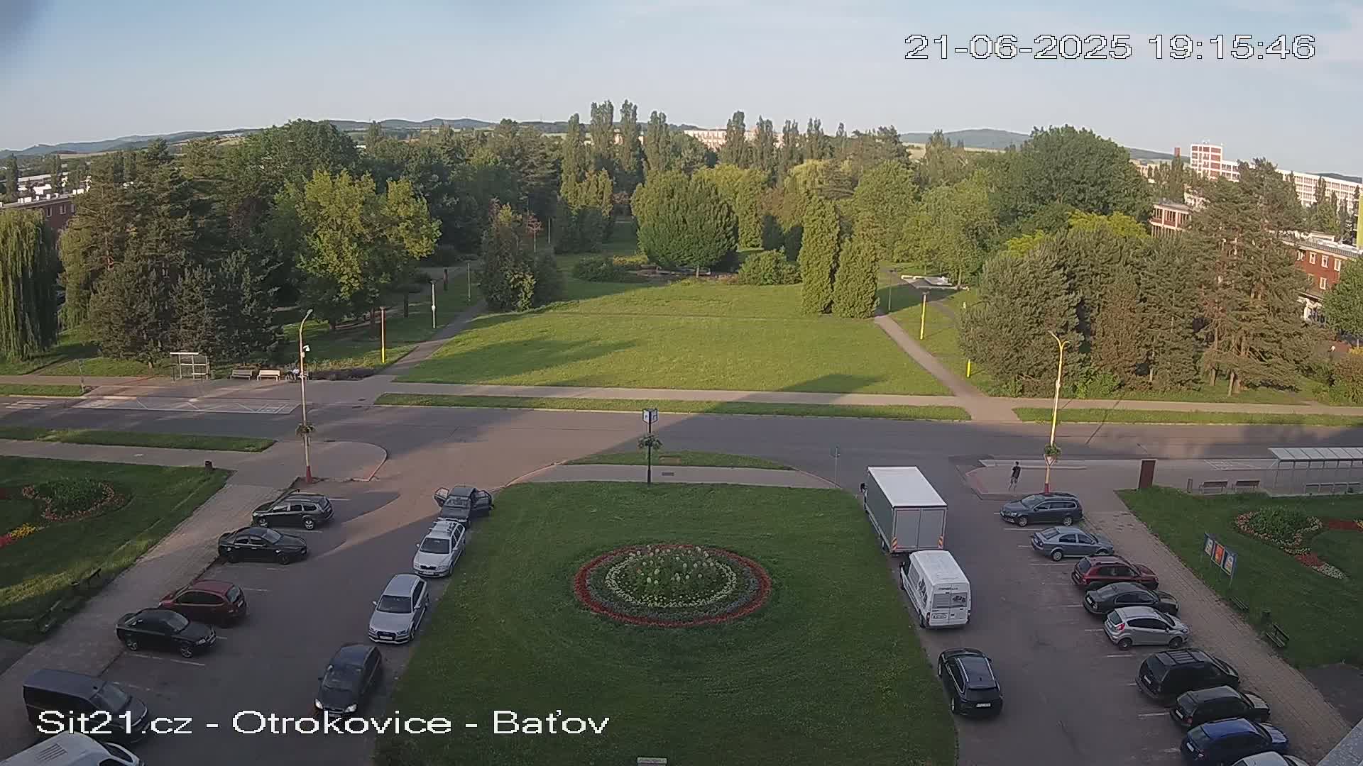 An aerial view shows a park with a large grassy area, several cars parked in a lot, and a truck and van stopped in the street on a clear evening.
