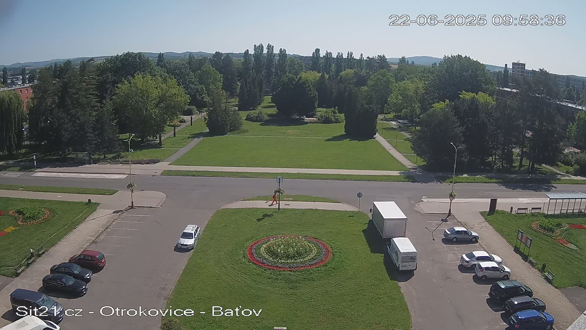 A sunny day reveals a high-angle view of a park with a central grassy area, surrounded by trees and roads, with cars parked in a lot and a few people walking.