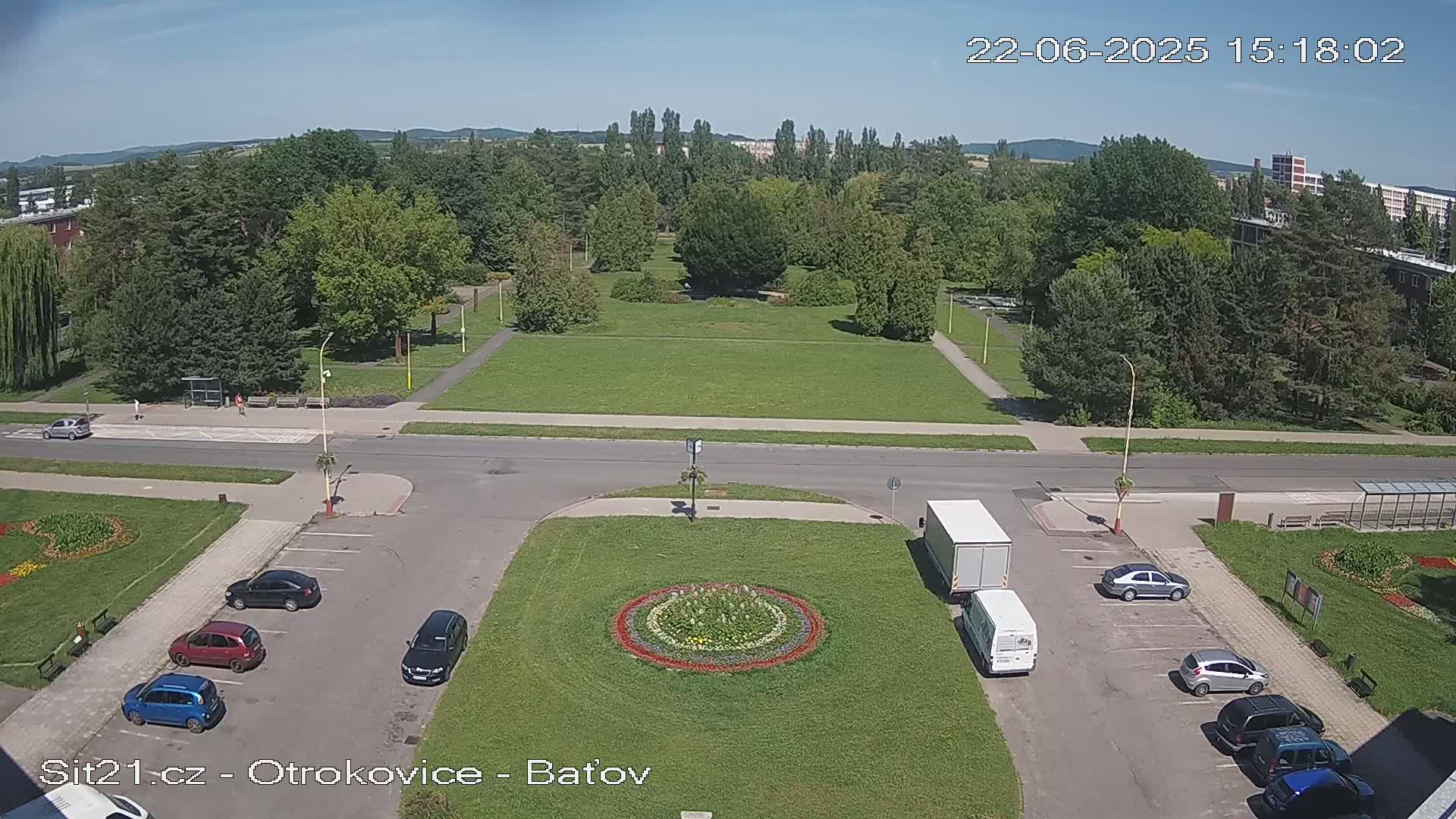 A sunny overhead view of a park and surrounding area with several cars parked in a lot, and two trucks, one of which is parked.