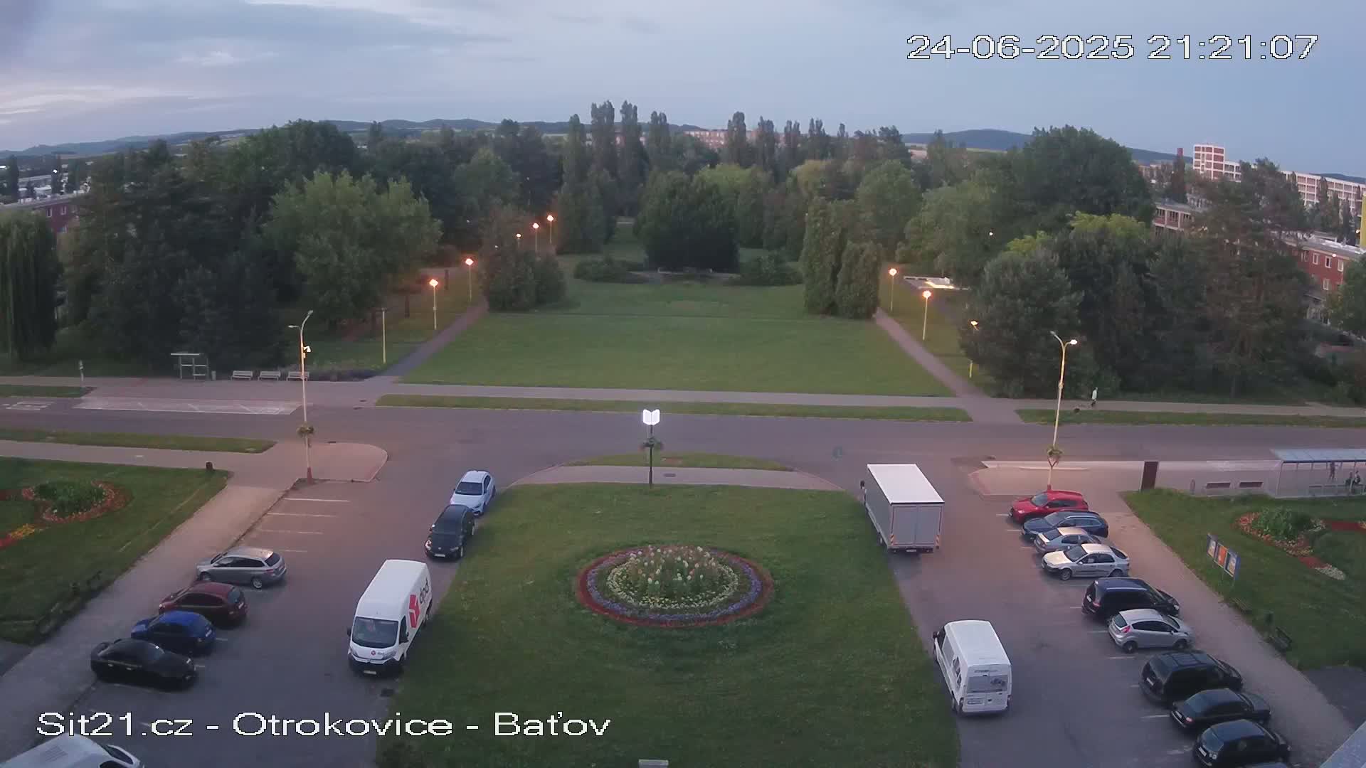 An aerial view of a town square at dusk, showing a grassy area with a circular flower bed in the center, surrounded by parked cars and vans, under a mostly clear sky.