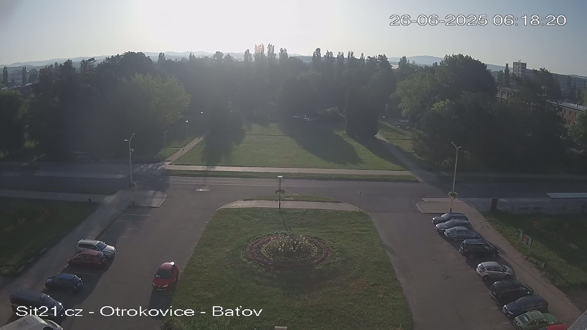 An aerial view of a town square at dusk, showing a grassy area with a circular flower bed in the center, surrounded by parked cars and vans, under a mostly clear sky.