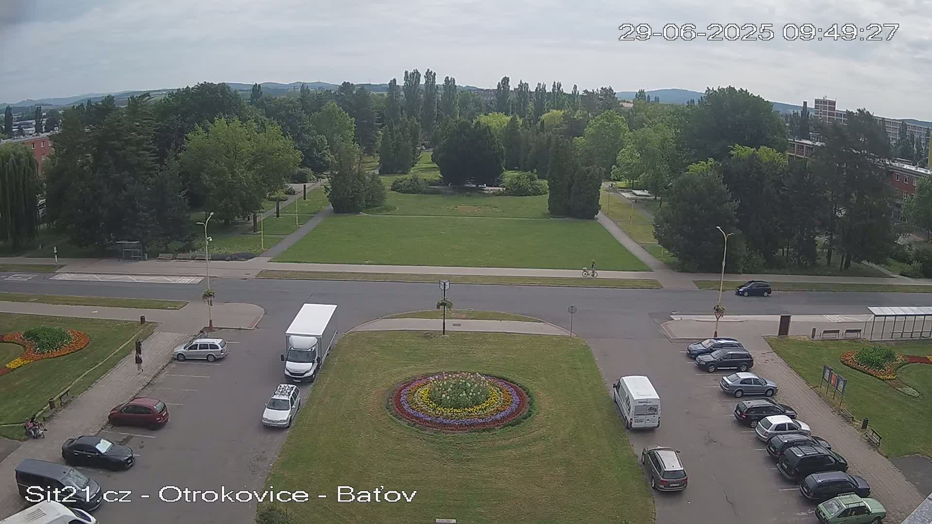 A high-angle view of a park with a large grassy area, surrounded by roads with parked cars and trucks, under a mostly cloudy sky.