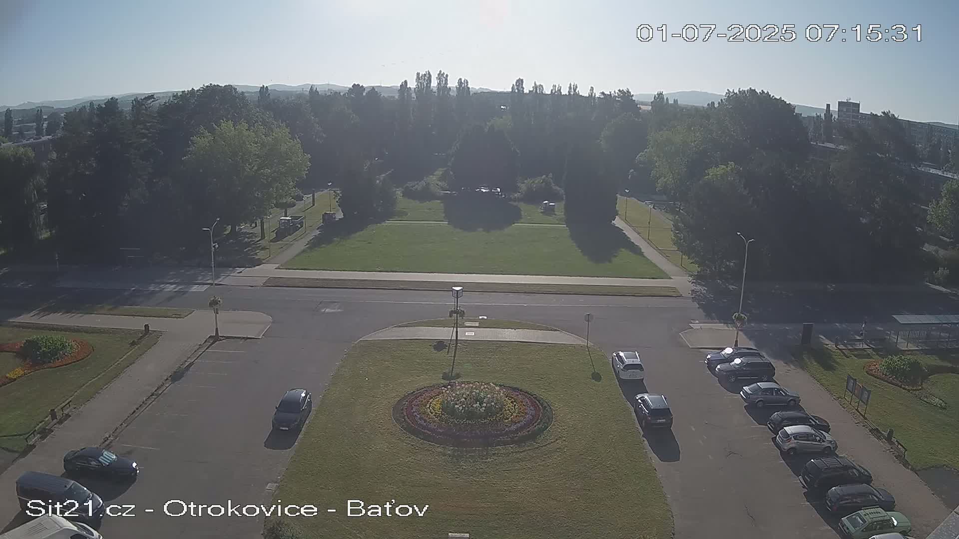 A high-angle, long shot shows a street intersection with a circular garden in the center, surrounded by a parking lot with several cars, and a park with many trees in the background on a hazy, sunny day.