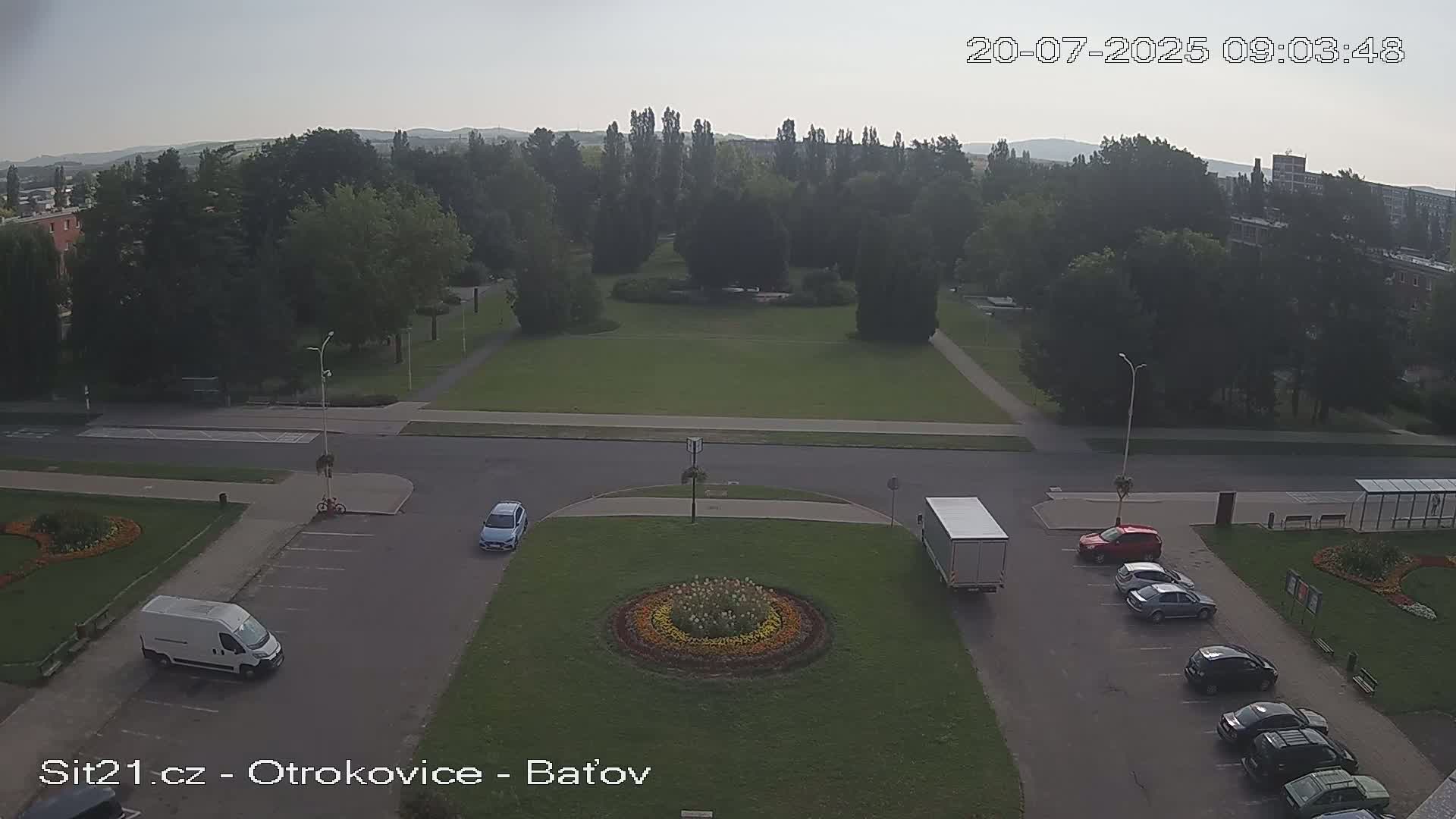 An aerial view of a park and parking lot shows several cars and a truck parked near a circular flower bed, on a hazy day.
