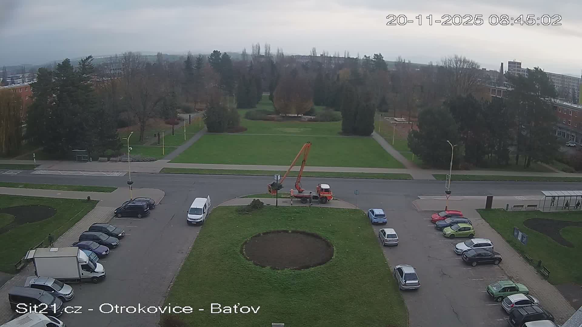 An overcast sky hangs over an urban area where a large park with deciduous and evergreen trees borders streets lined with parked cars, and an orange crane truck is positioned on a grassy median.