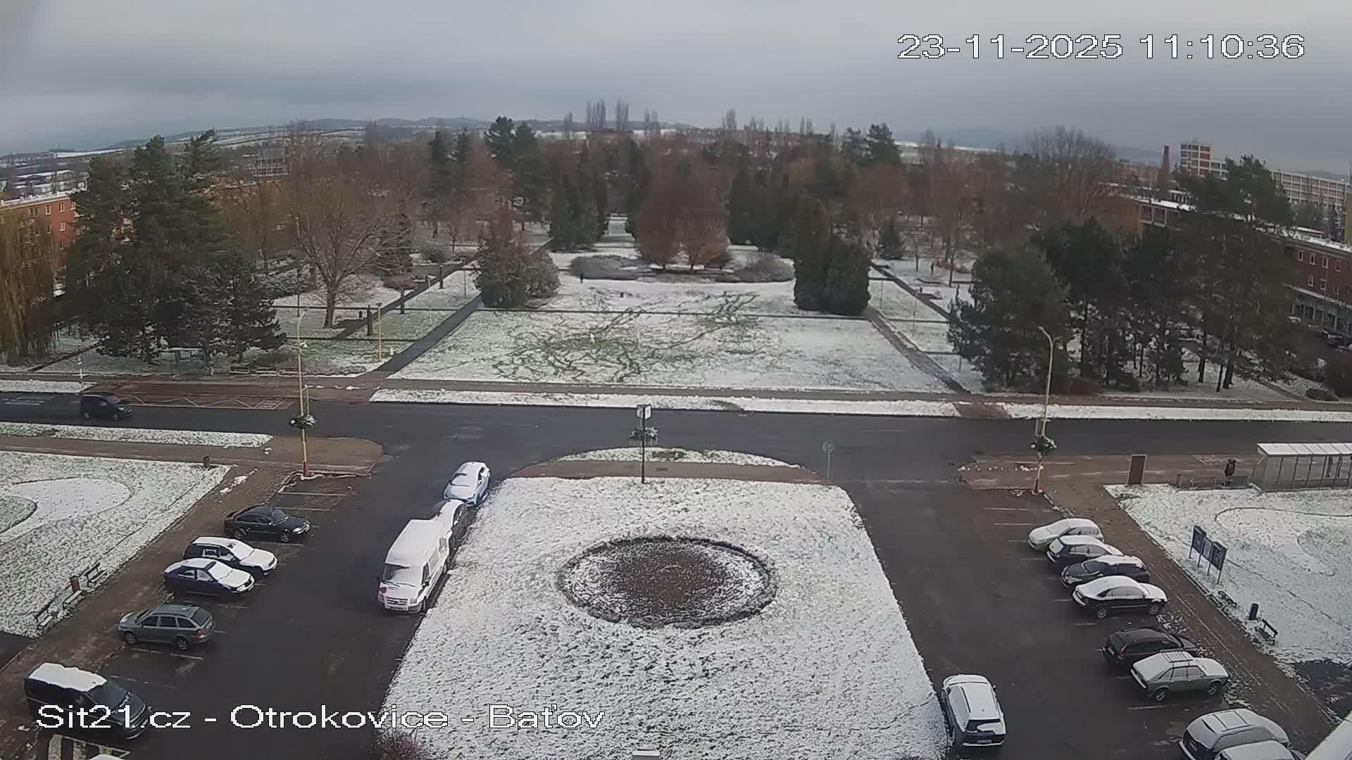 An overhead view captures a city street and park lightly dusted with snow under an overcast sky, with bare trees, parked cars, and distant buildings.