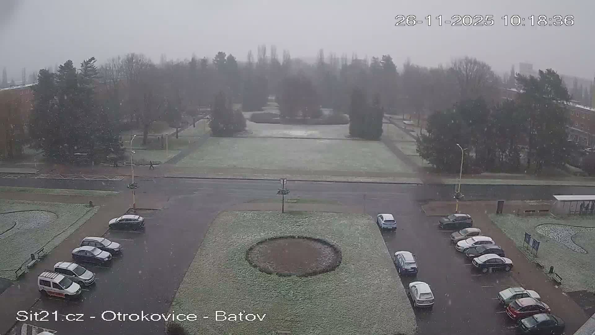 Light snow falls on an overcast winter day over a paved street with parked cars, bordering a frosted green park with trees, and a few pedestrians.