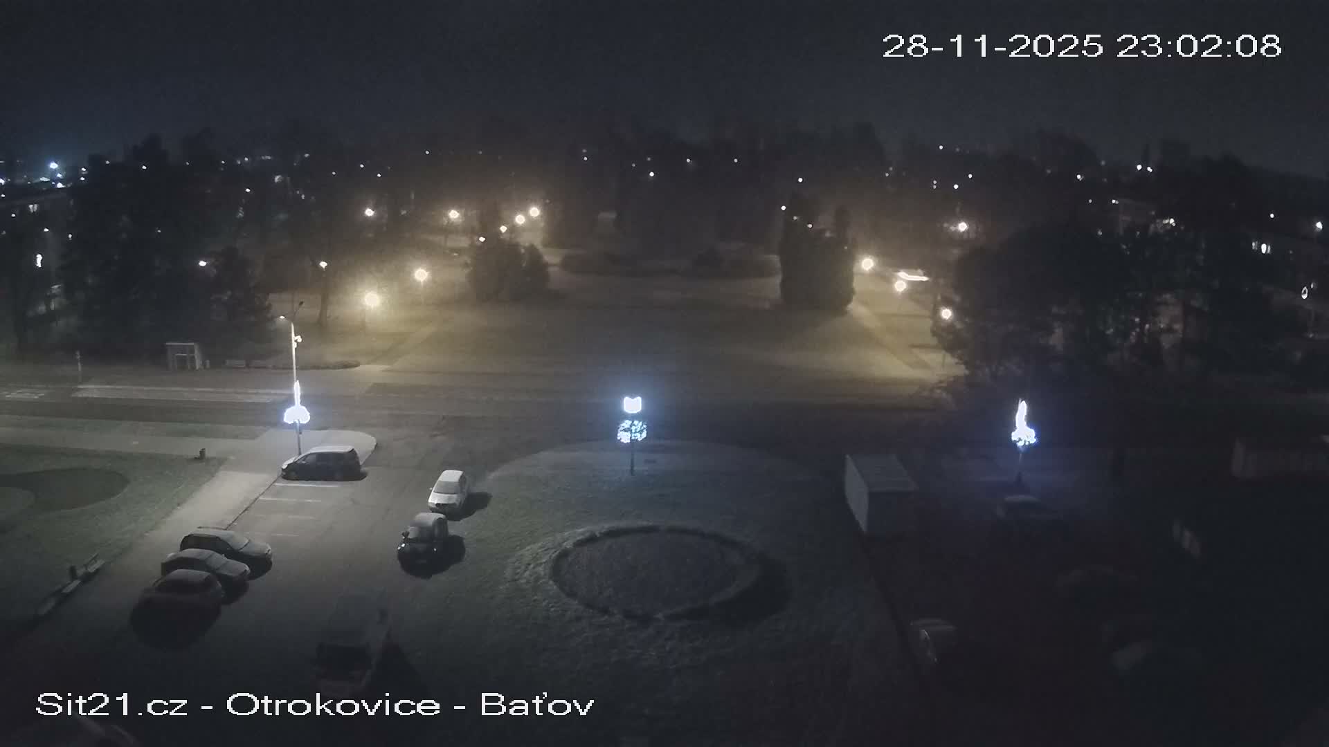 An elevated night-time view depicts a quiet urban area with parked cars, trees, and roads illuminated by streetlights, under clear yet somewhat damp or frosty conditions.
